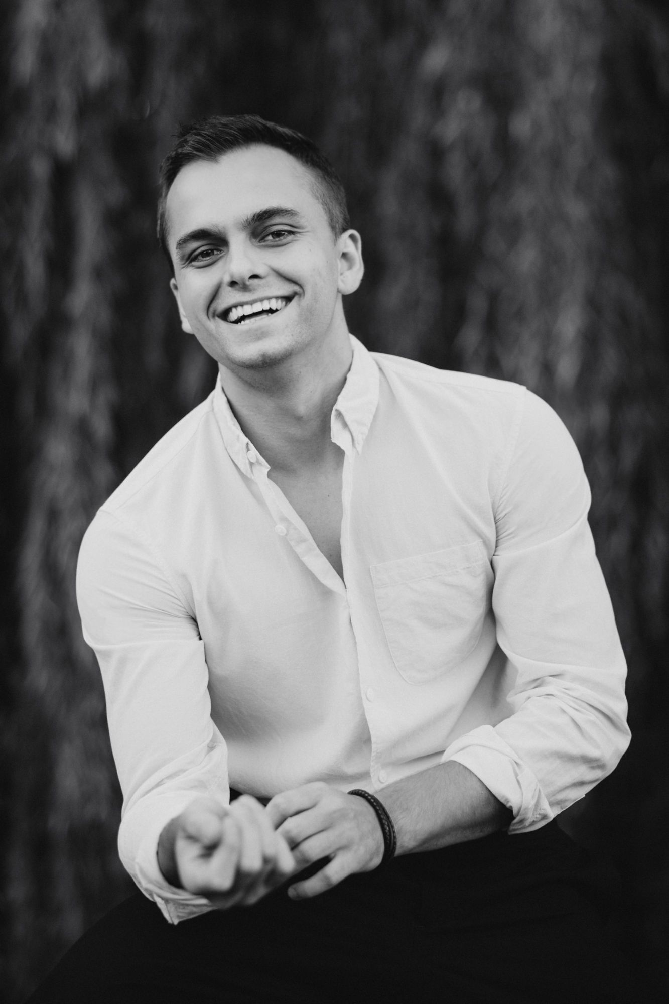 Black and white portrait photograph of person in white button-down shirt smiling warmly against blurred background.