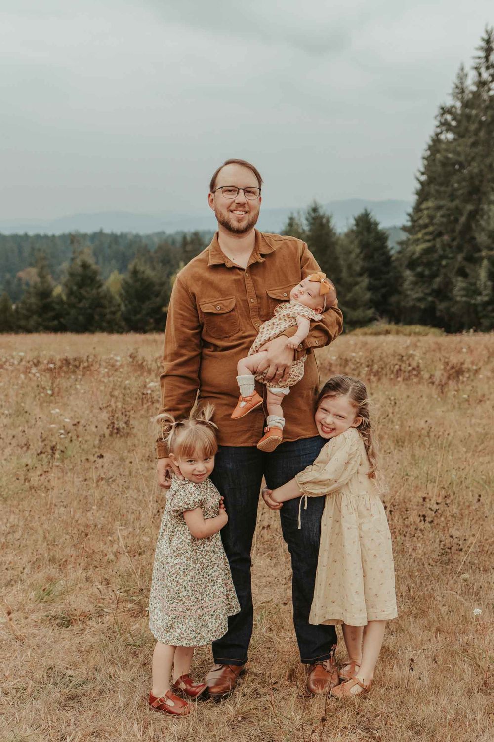 three little girls snuggling their dad in a big open field all wearing a neutral color pallette