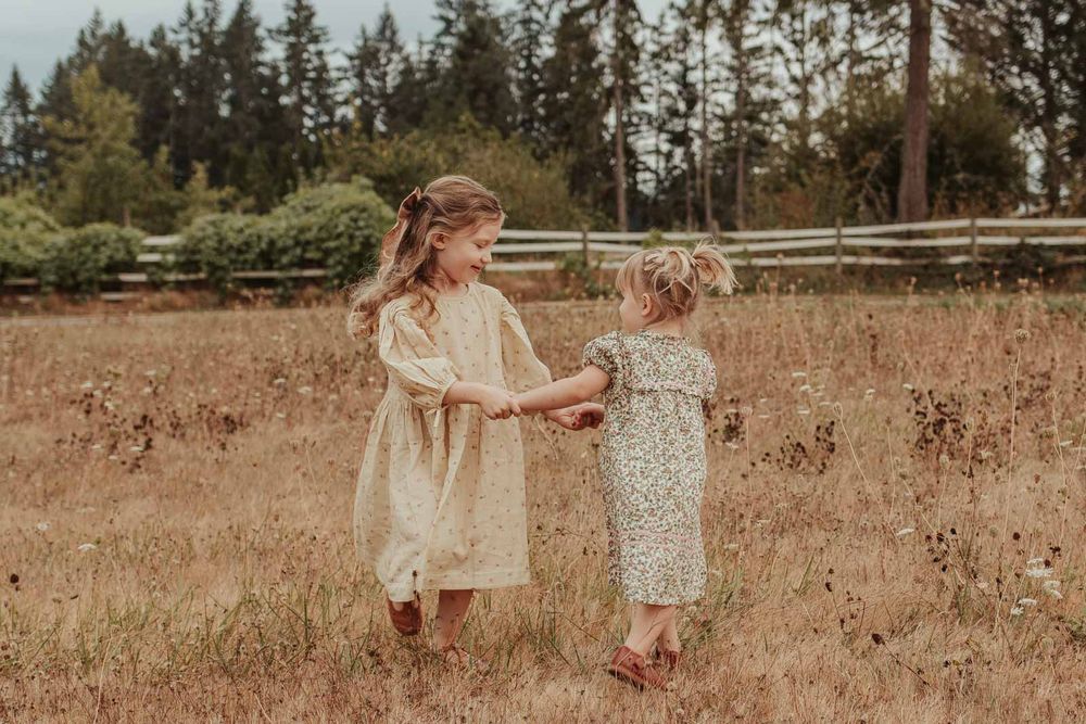 two little sisters holding hands and spinning in a grassy field wearing floral dresses and bows