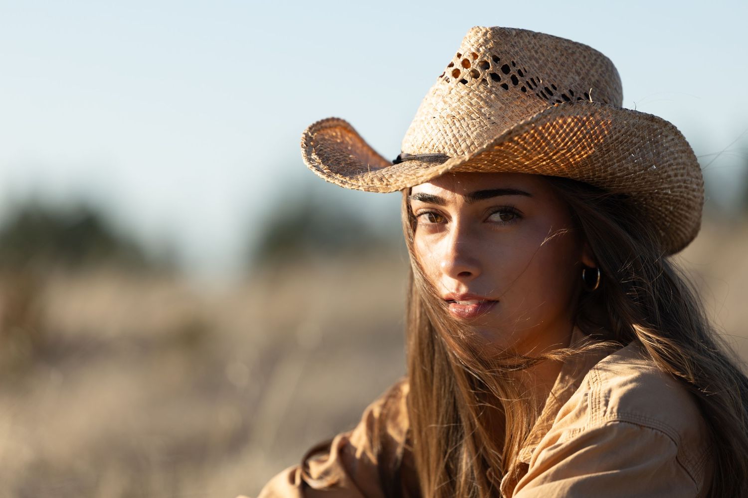 A woman wearing a cowboy hat poses outdoors against a sunny desert landscape with a natural bokeh effect.
