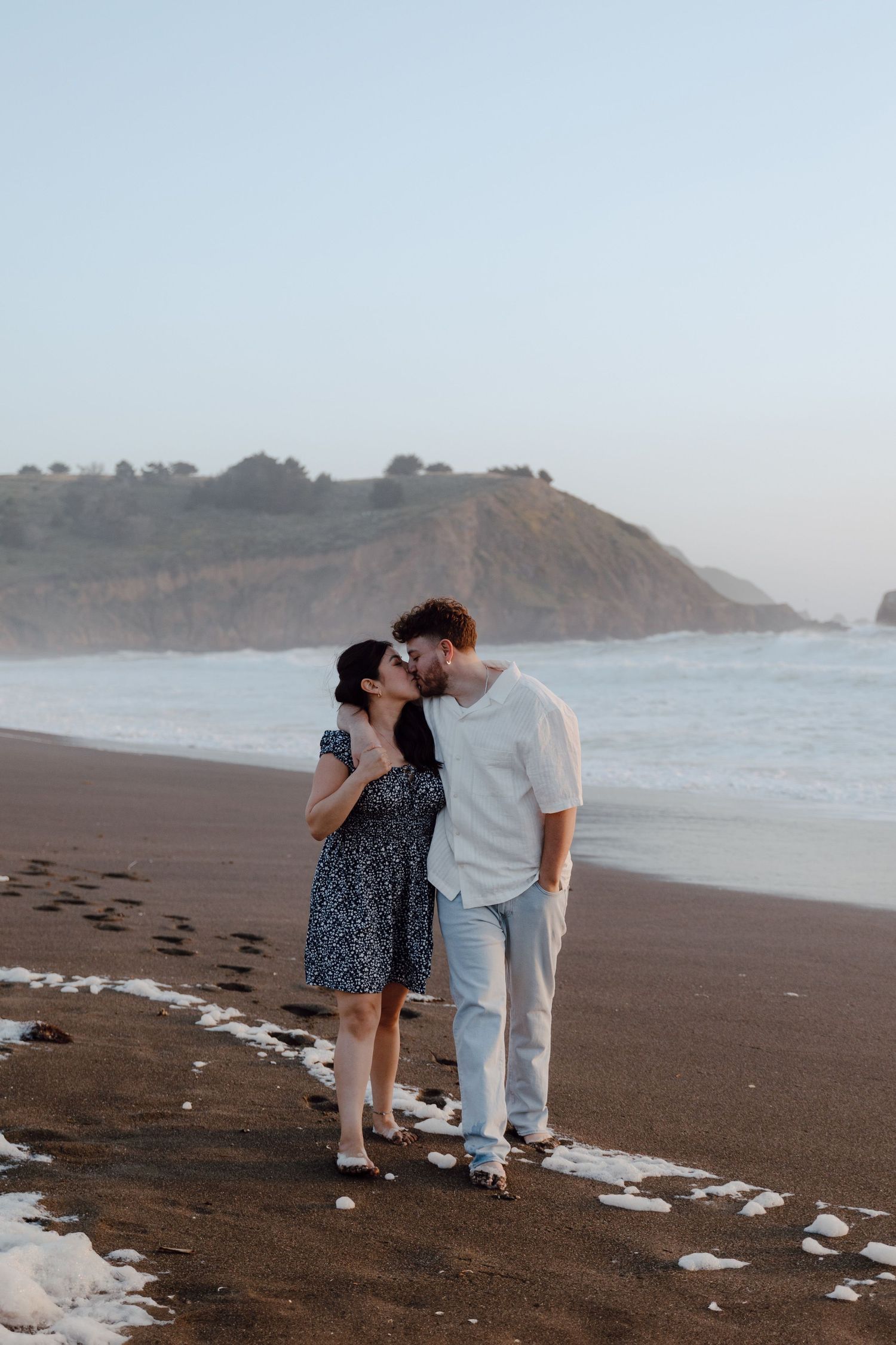 Couple sharing intimate moment on foggy beach with coastal cliffs in background.