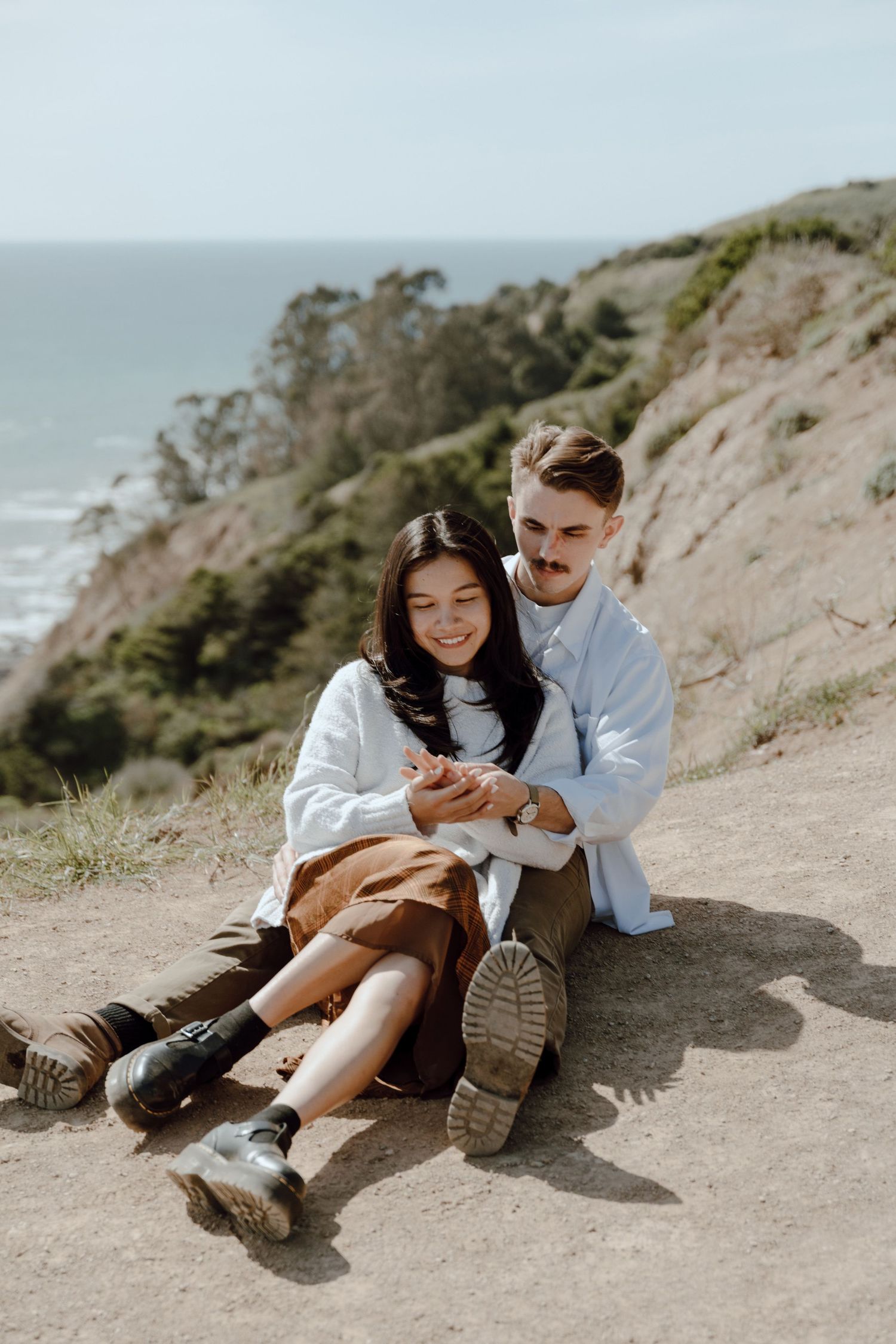 Couple sitting on coastal cliff path overlooking ocean on sunny day.
