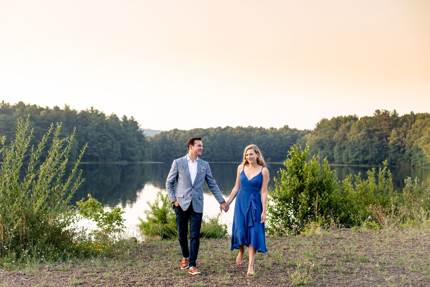 A couple walking hand in hand near a serene lake during golden hour.