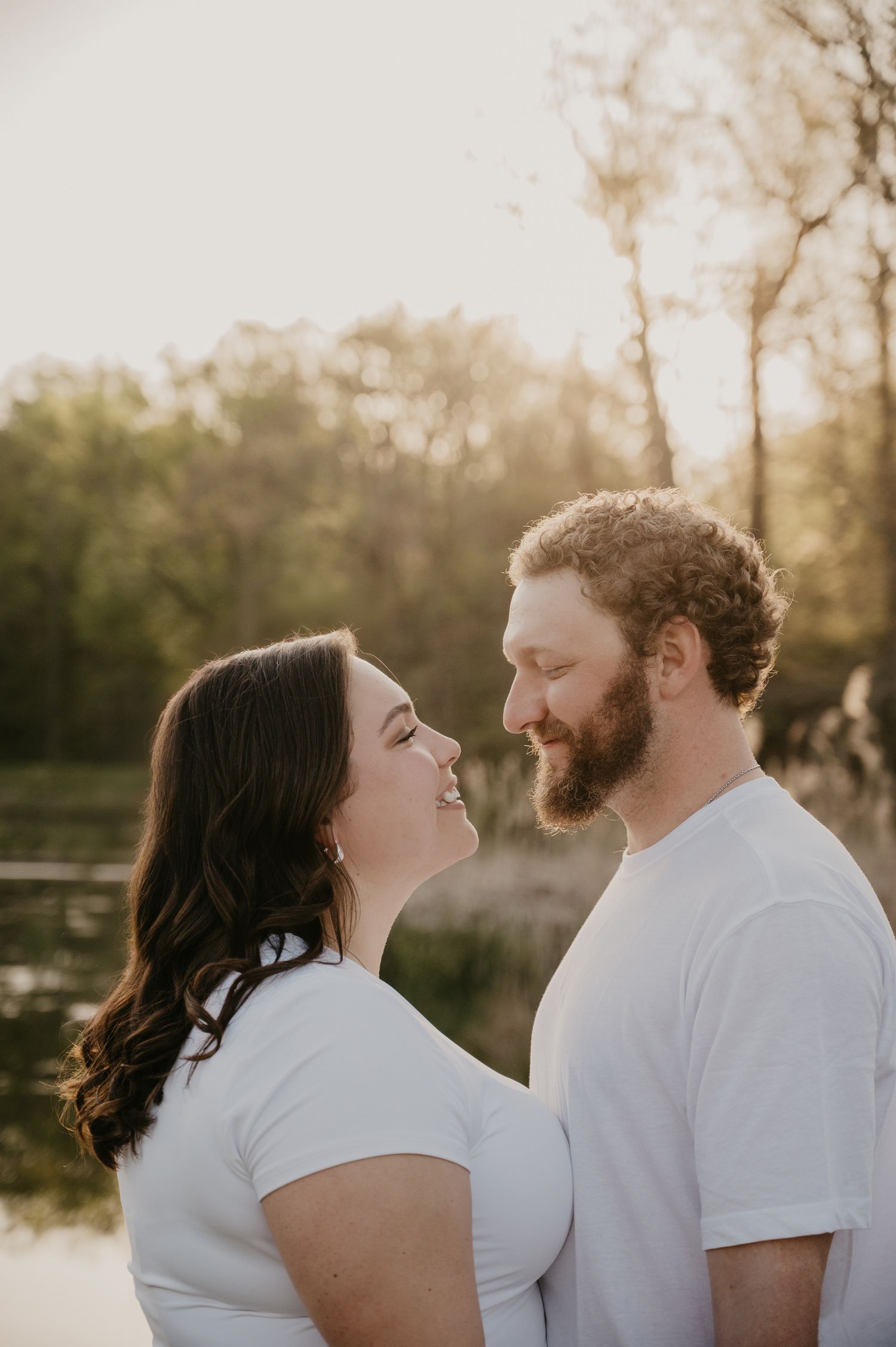 A smiling couple in matching white shirts shares a romantic moment by a sunlit lake.
