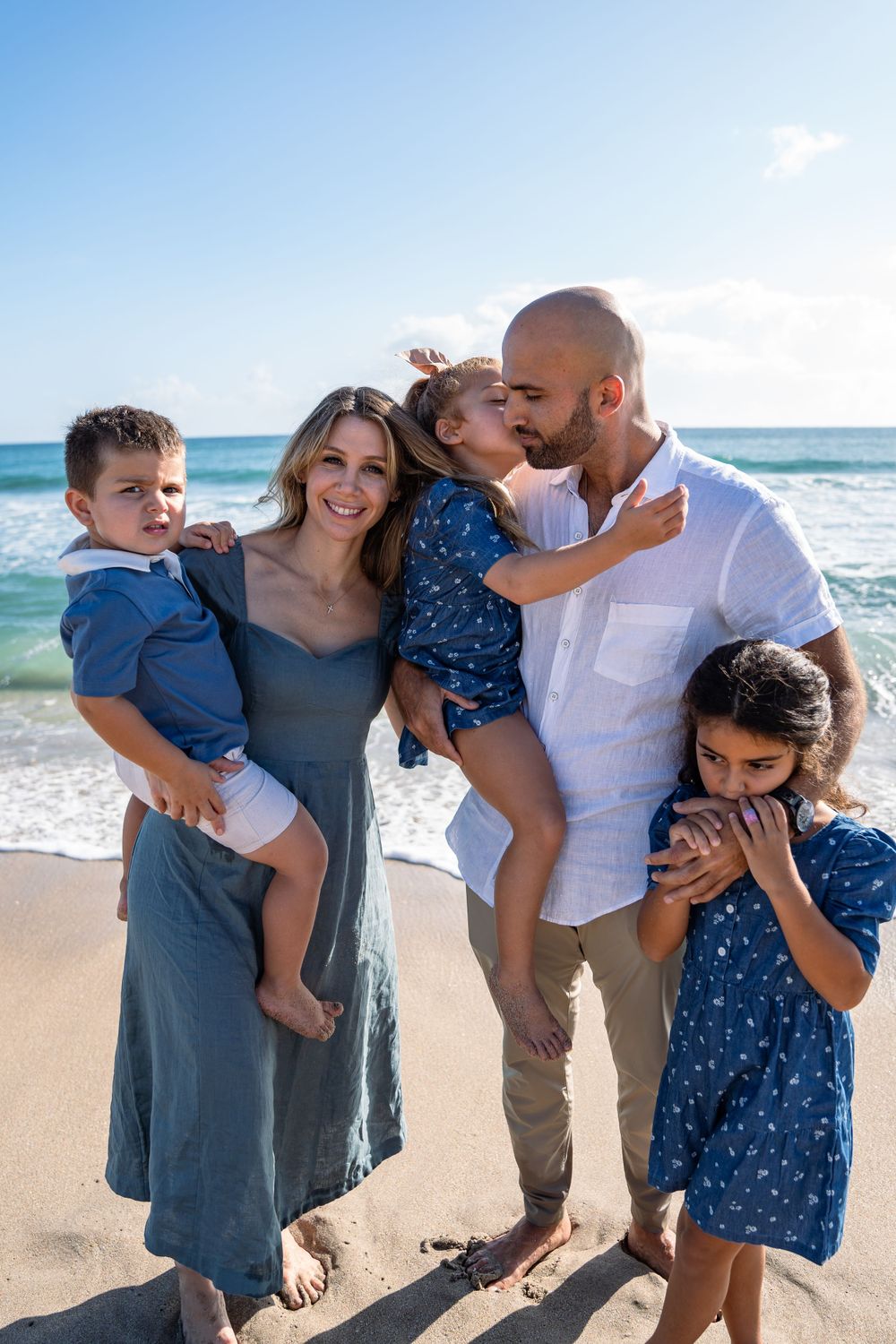 Affectionate family moment captured on a sunny beach with ocean waves in background.
