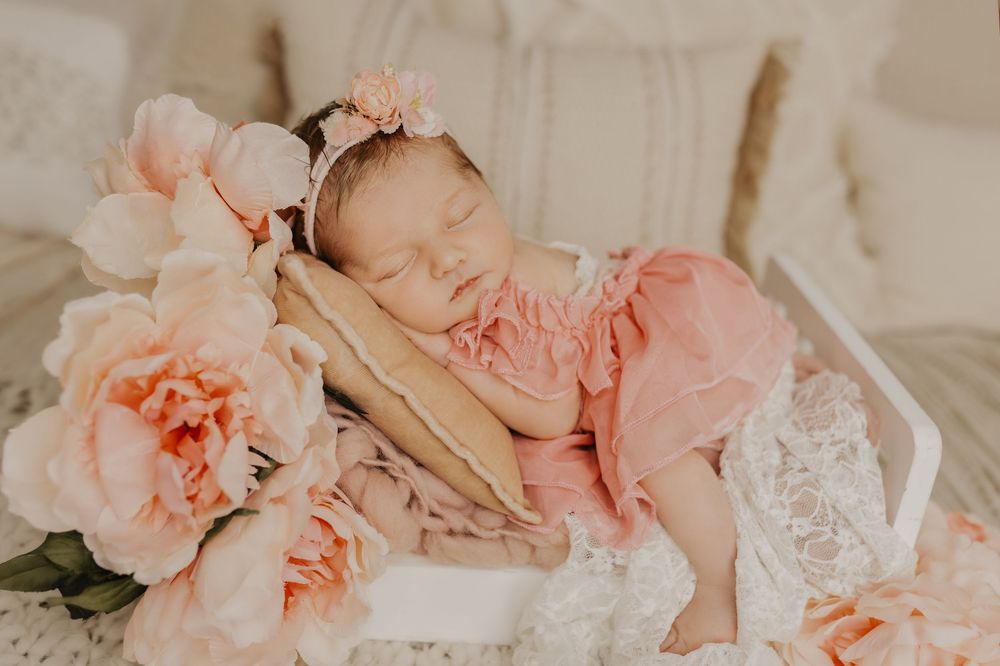 Newborn baby girl sleeping peacefully in pink ruffle outfit surrounded by blush peony flowers.