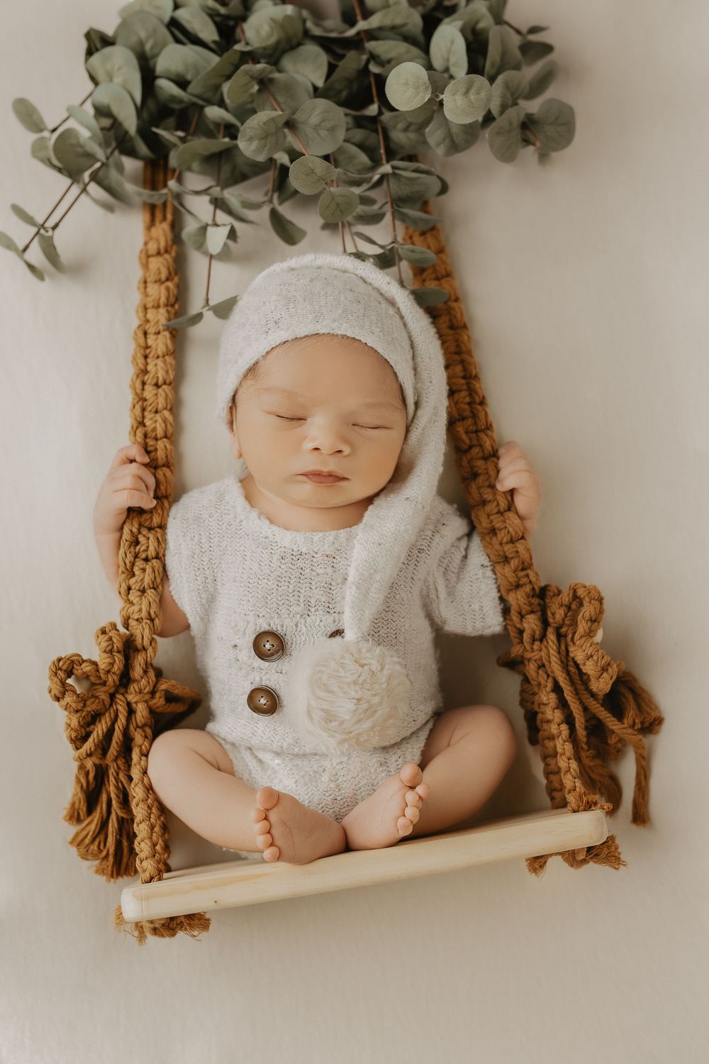Sleeping newborn baby in white knit romper and bonnet posed on a rustic macrame swing with eucalyptus.