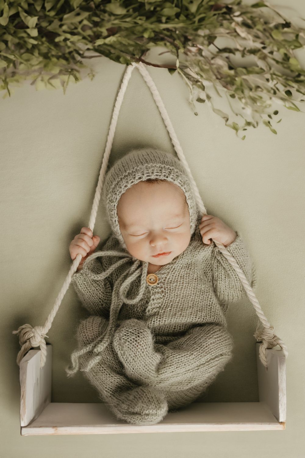 Sleeping infant wearing gray knit outfit lying on white swing prop with greenery accents.