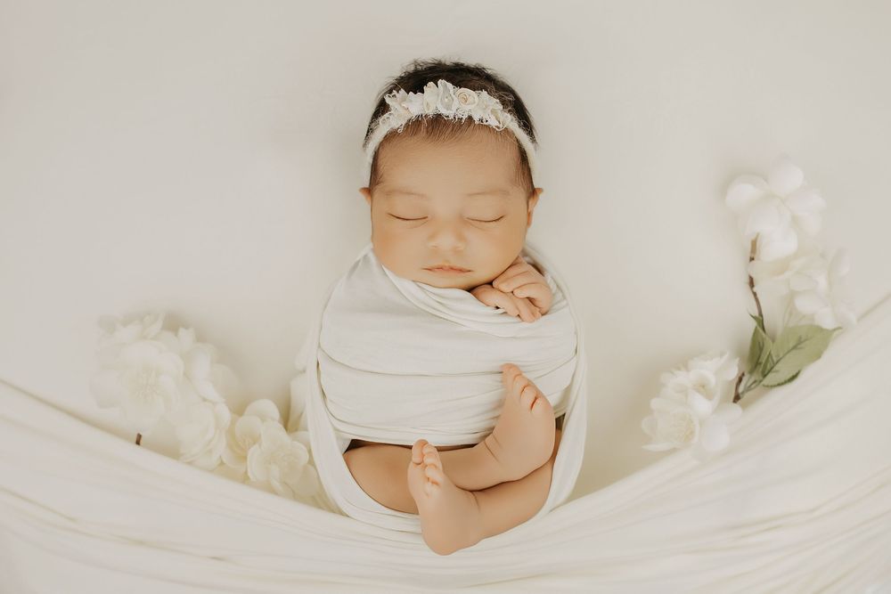Sleeping newborn baby wrapped in white swaddle with floral headband surrounded by white flowers.