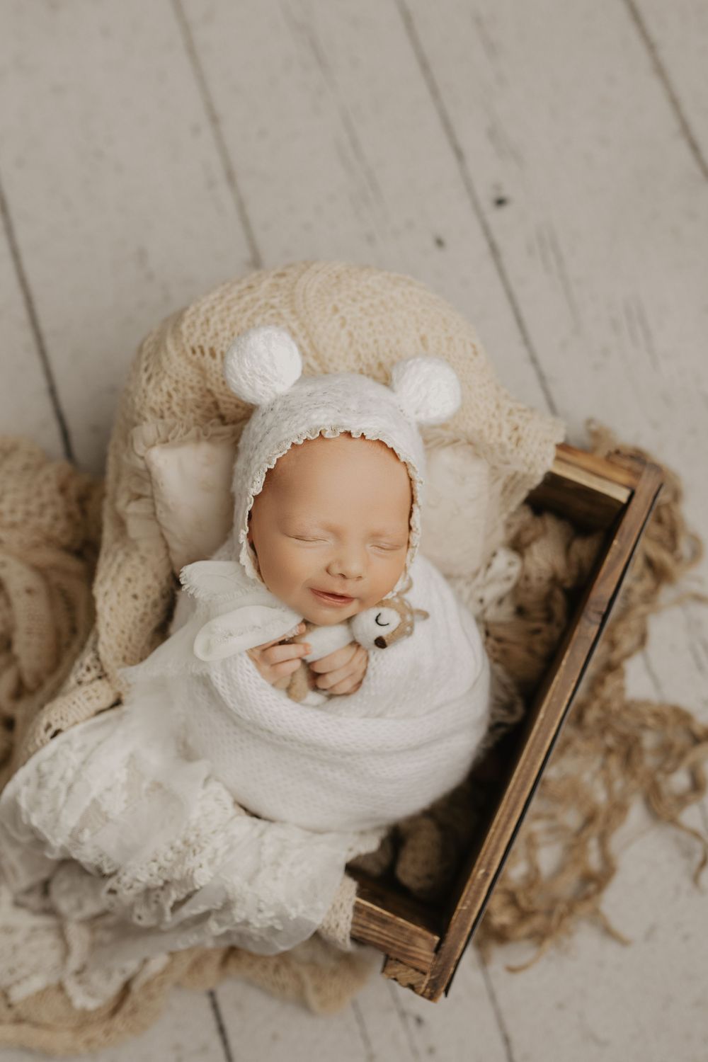 Sleeping newborn in white knit bear bonnet on rustic wooden floor.