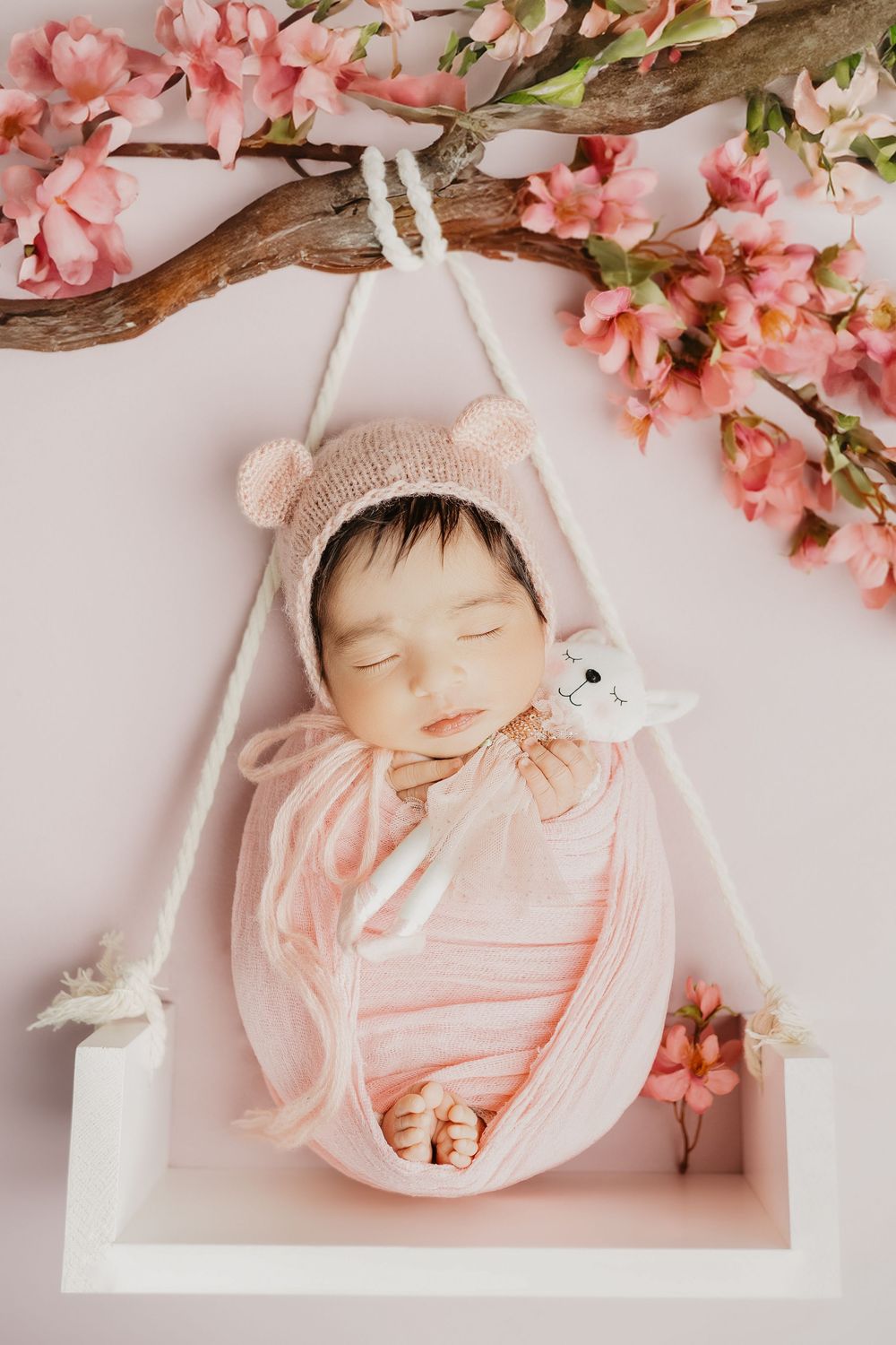 Sleeping baby in pink outfit under cherry blossom branches on white swing.