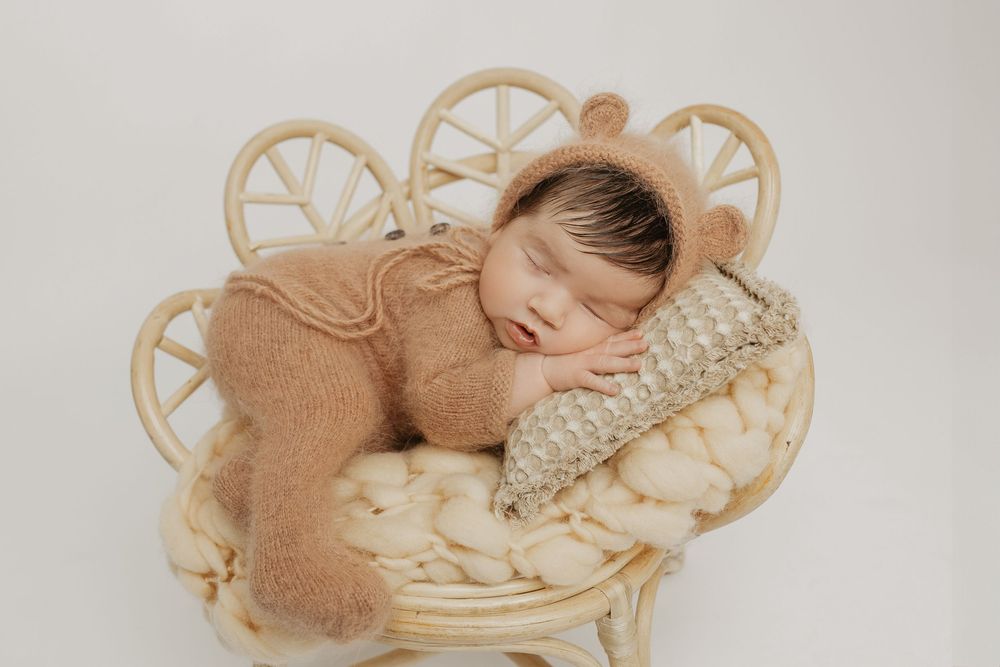 Sleeping newborn posed in antique wicker basket with peach-colored accessories.