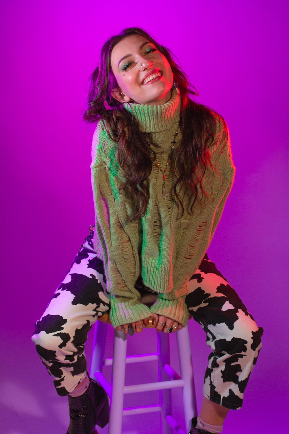 Portrait of someone in knit sweater and cow print pants sitting on stool with purple lighting.