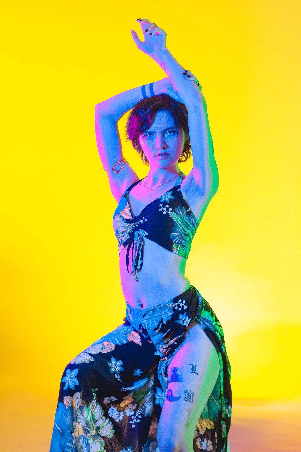Dancer in floral two-piece outfit striking elegant pose against yellow background with colorful lighting.