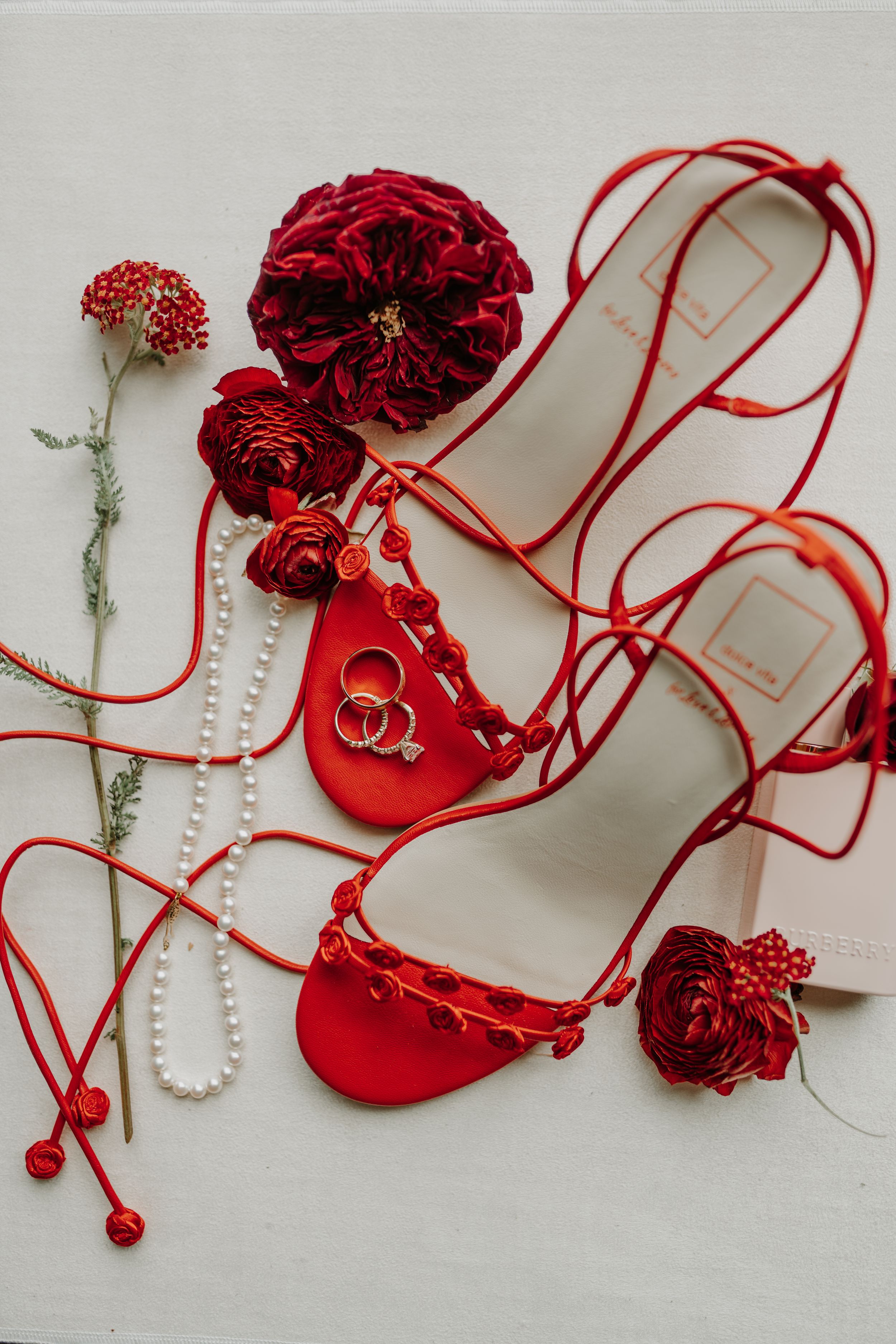 Red wedding accessories including floral shoes, rings, and roses arranged on white background.