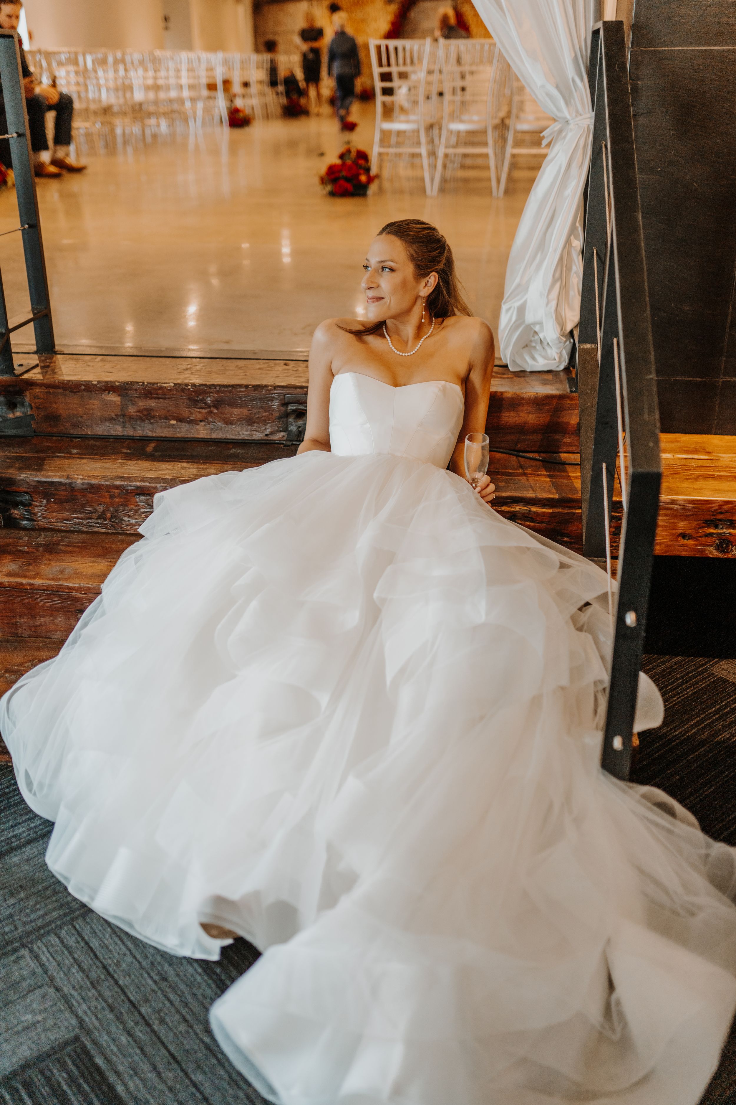 Beautiful bride in a flowing white ballgown dress sits gracefully on wooden steps at wedding venue.