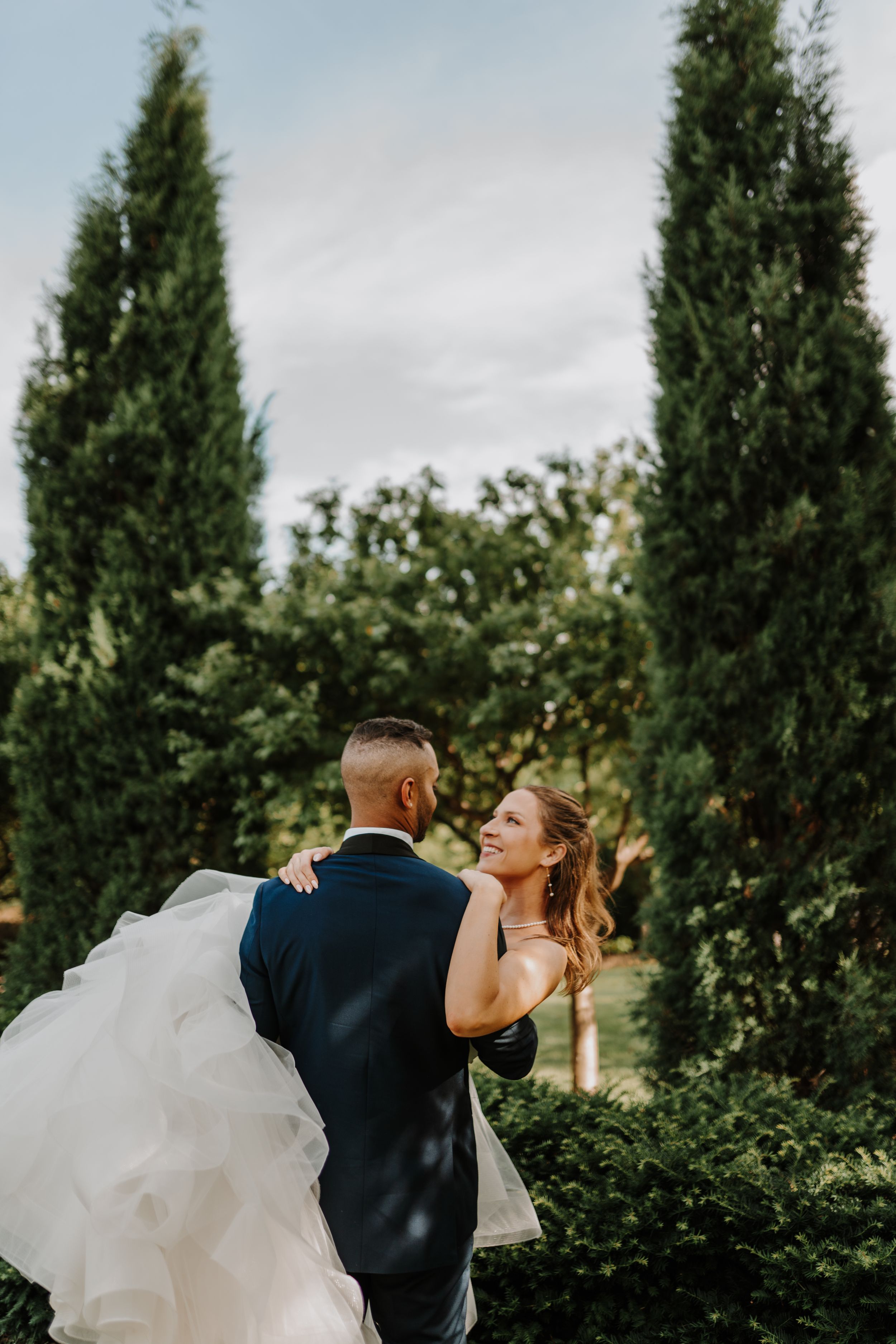 Romantic outdoor wedding photo with newlyweds embracing among tall cypress trees.
