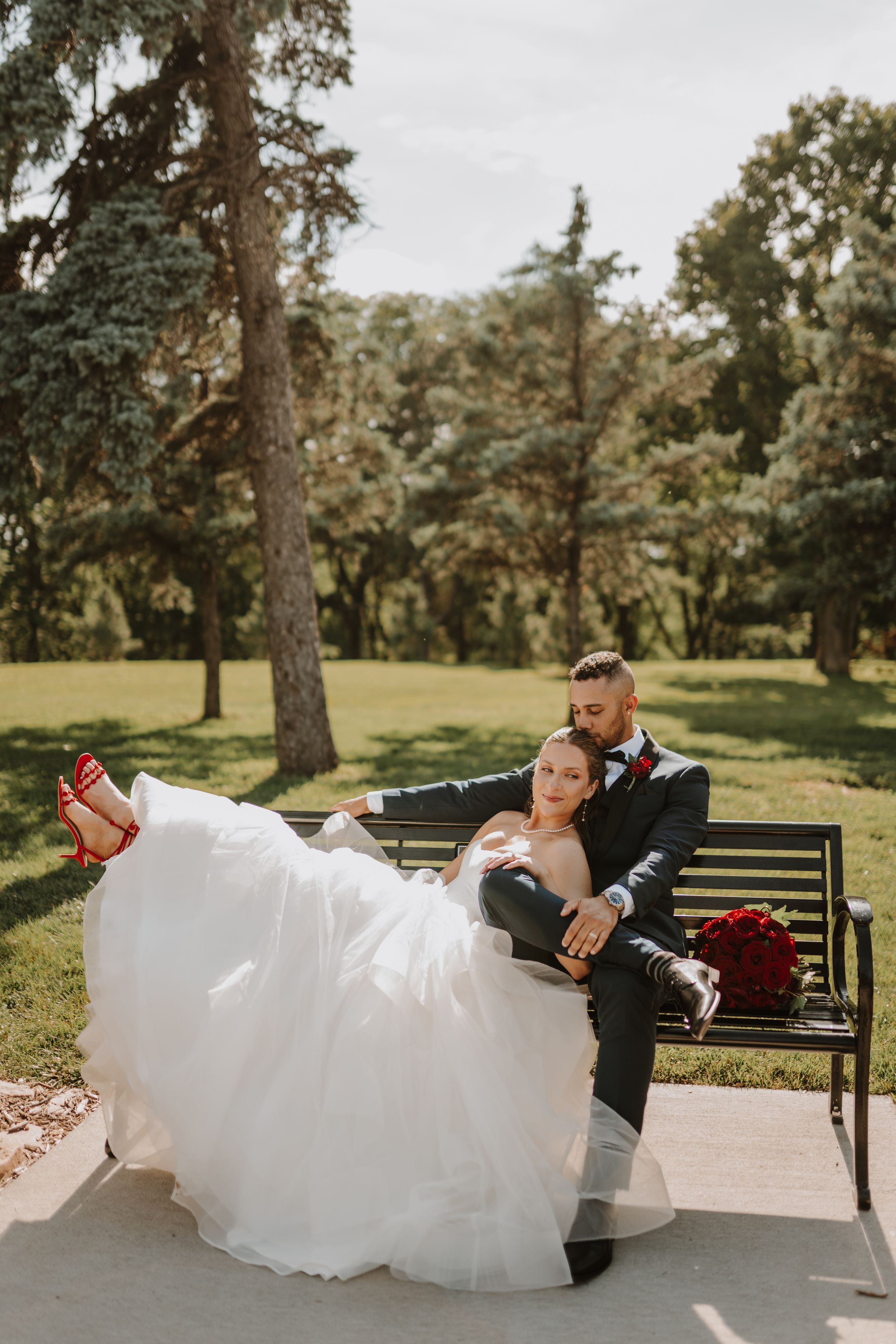 Newlyweds relax on park bench with bride's dress elegantly draped.