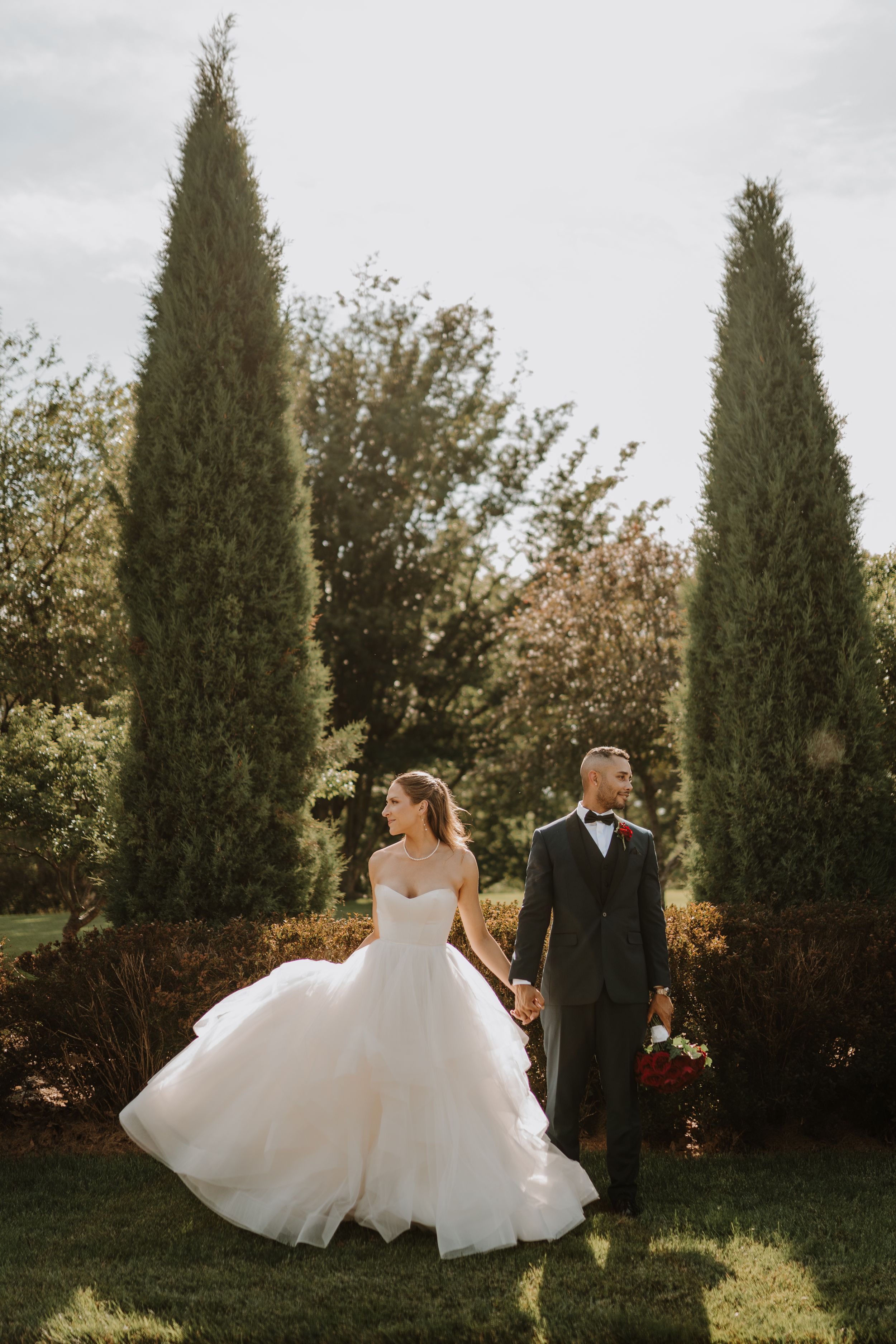 A couple in wedding attire poses between tall cypress trees in a garden setting during golden hour.