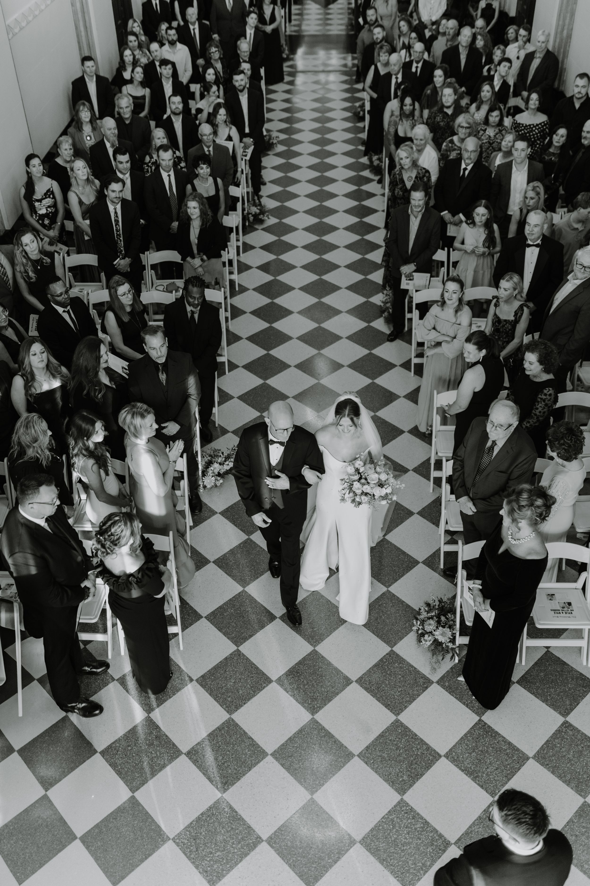 Aerial black and white photo of wedding ceremony in grand hall with checkered floor.