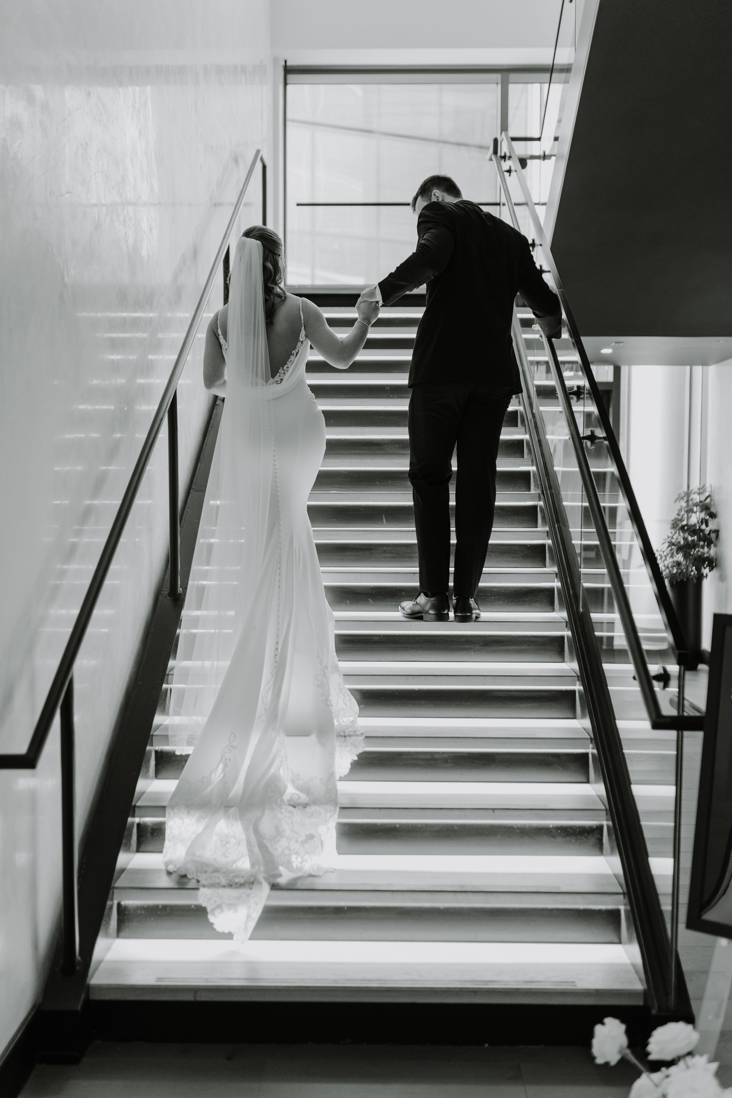 Black and white photo of couple ascending modern staircase in wedding attire.