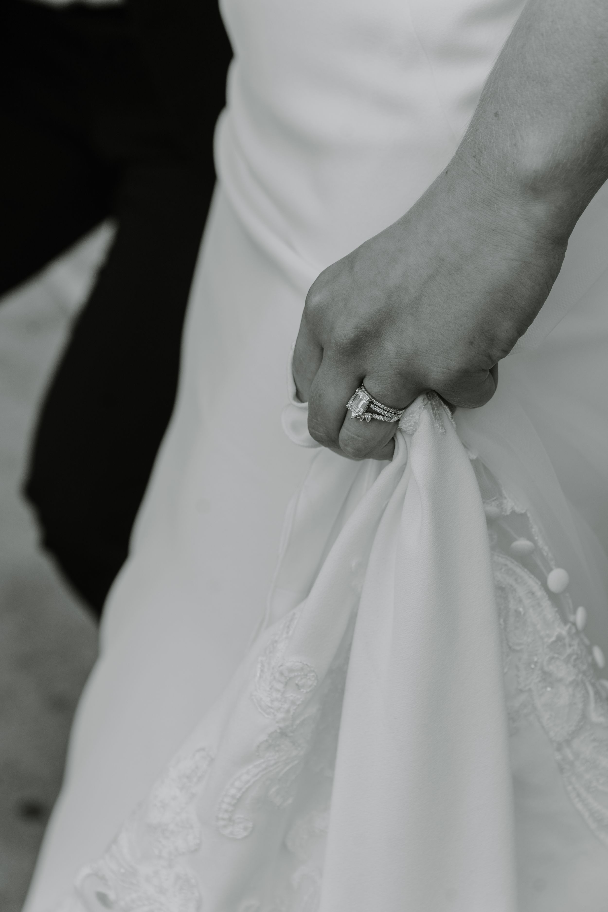 Close-up black and white photo of bride's hand holding wedding dress, showing ring.