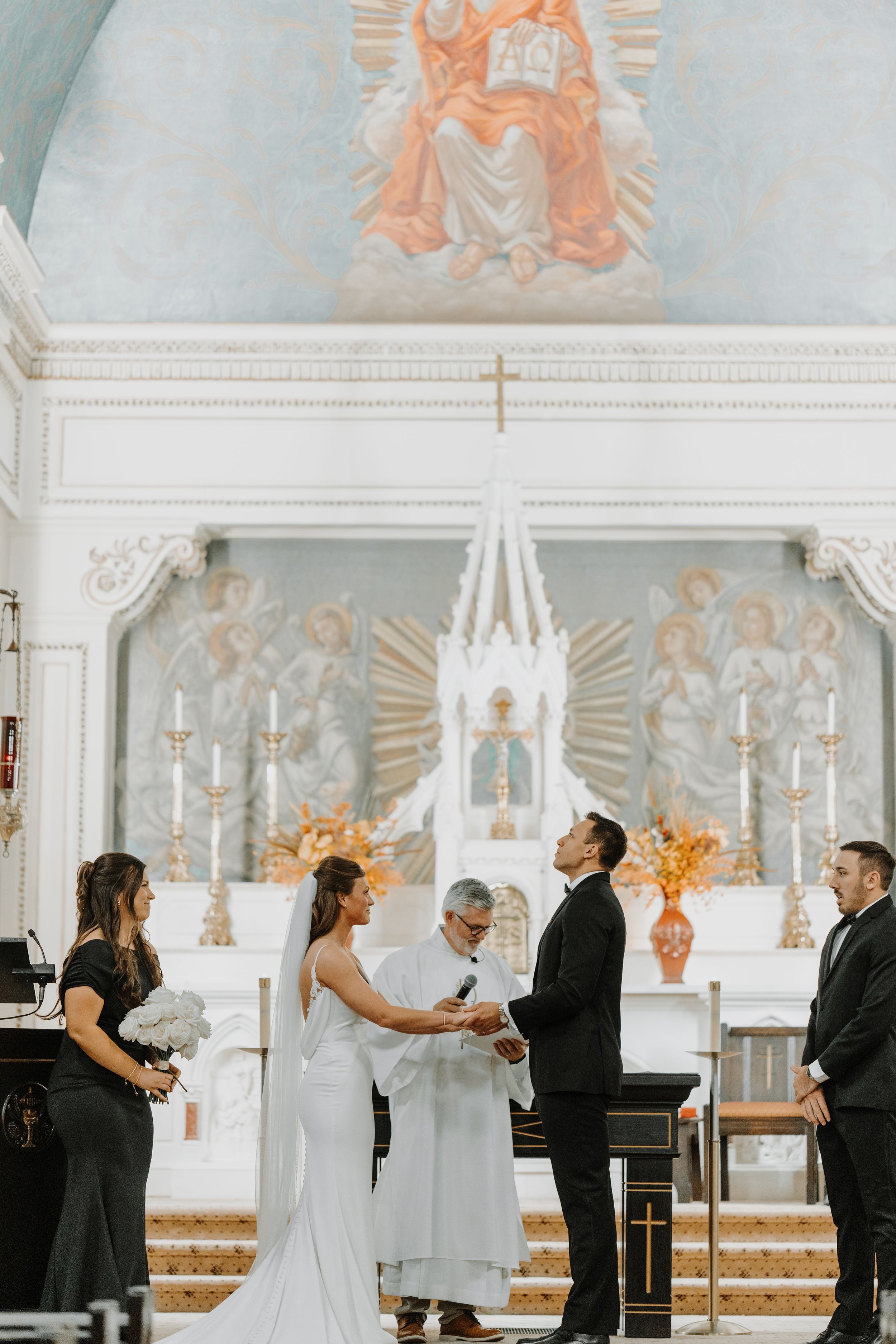 Wedding ceremony scene in an elegant white church with ornate wall paintings and golden accents.