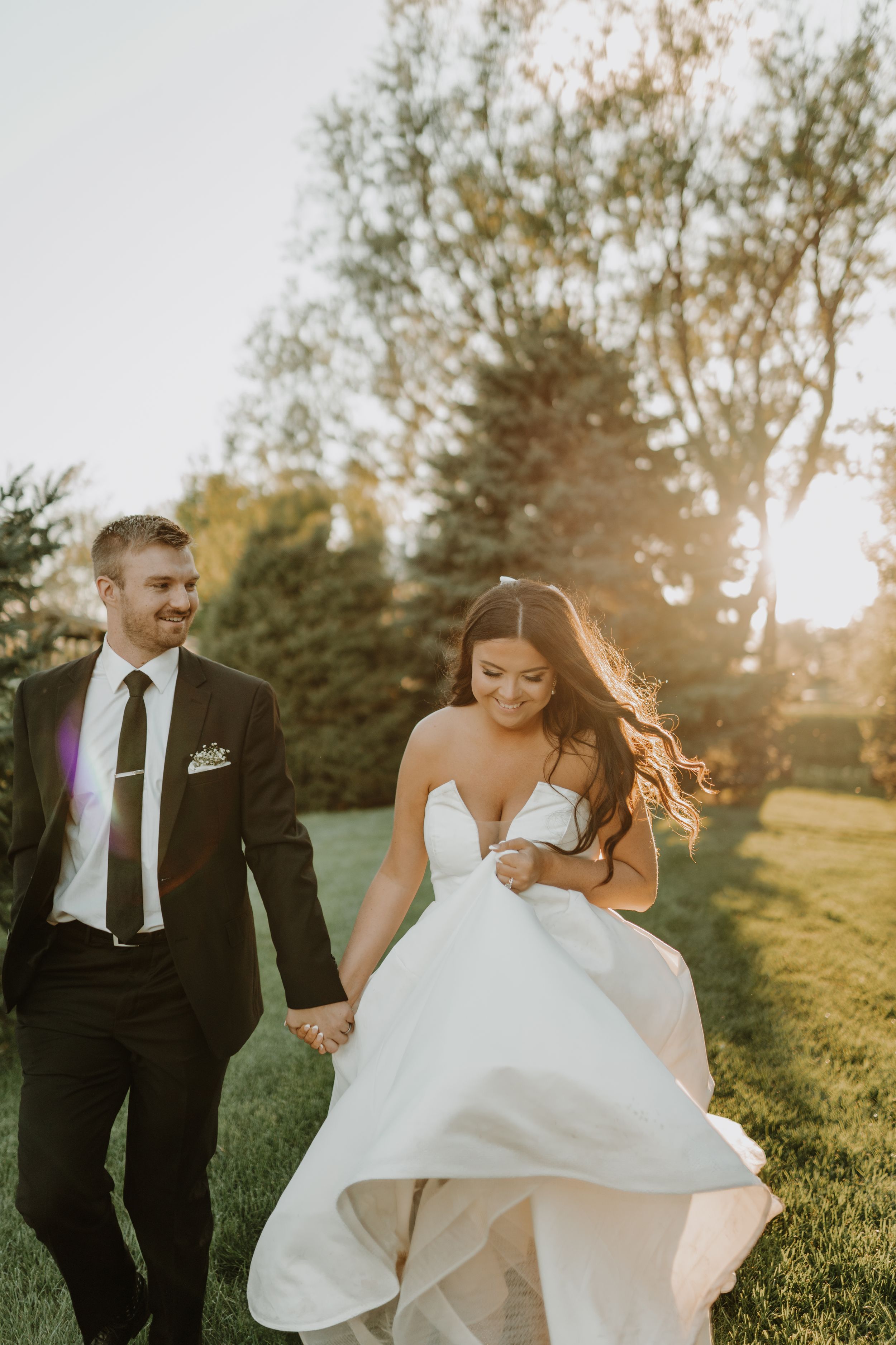 Newlyweds walk hand in hand through a sunlit garden as the bride's white dress flows in the golden hour light.