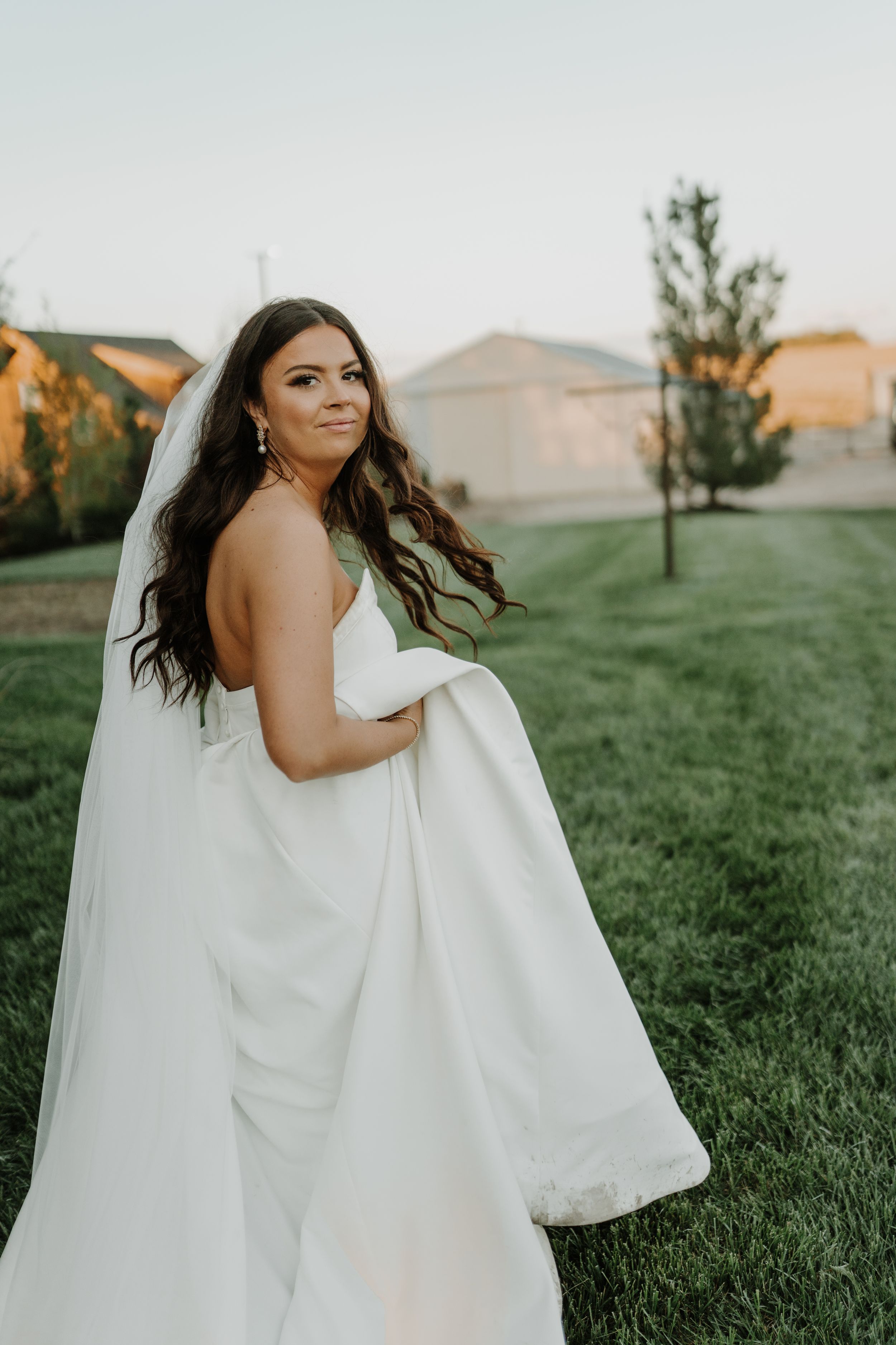 Bride in flowing white dress poses alone in garden with windswept hair at sunset.