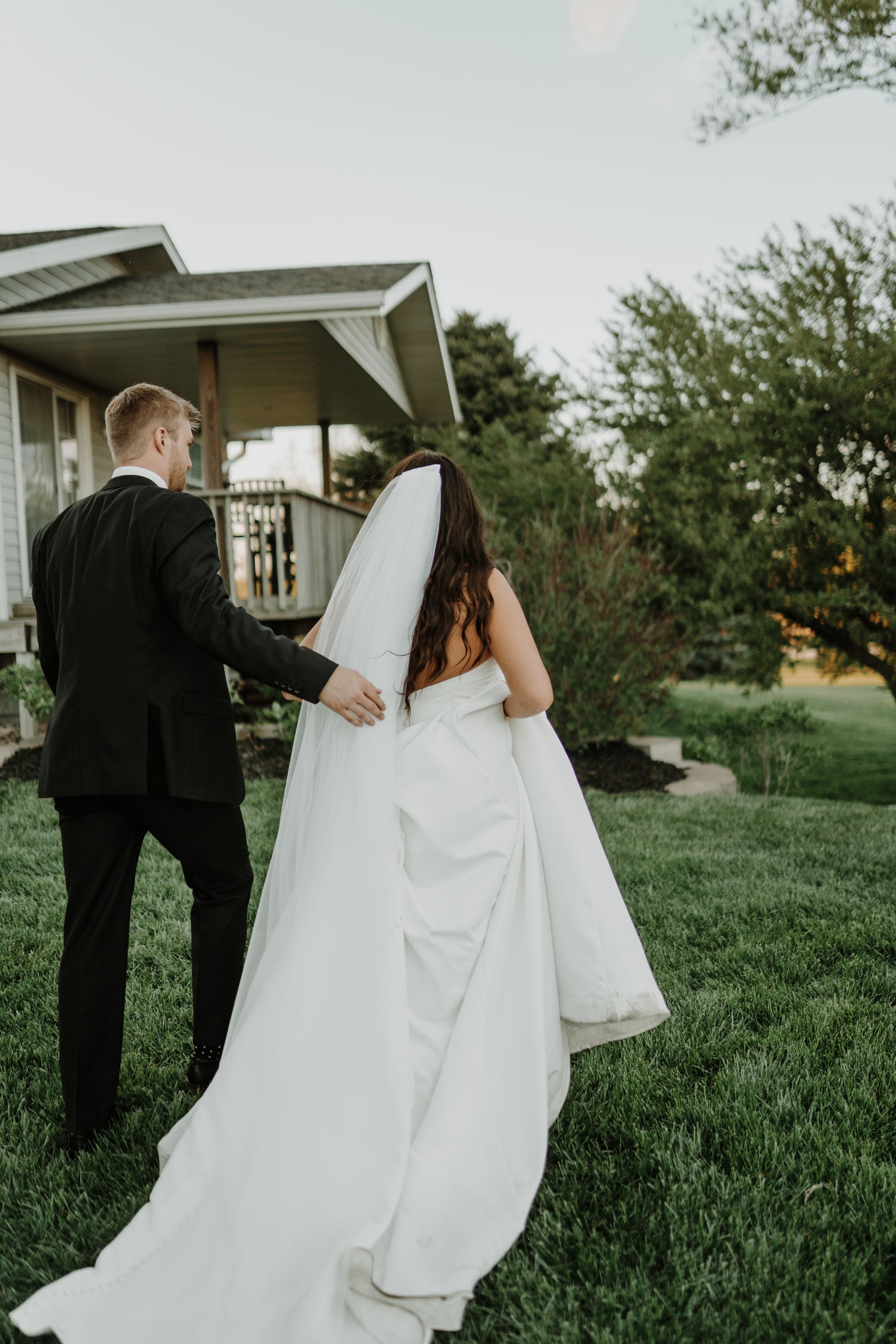 Wedding couple walking up garden path towards house as bride's dress trails behind.