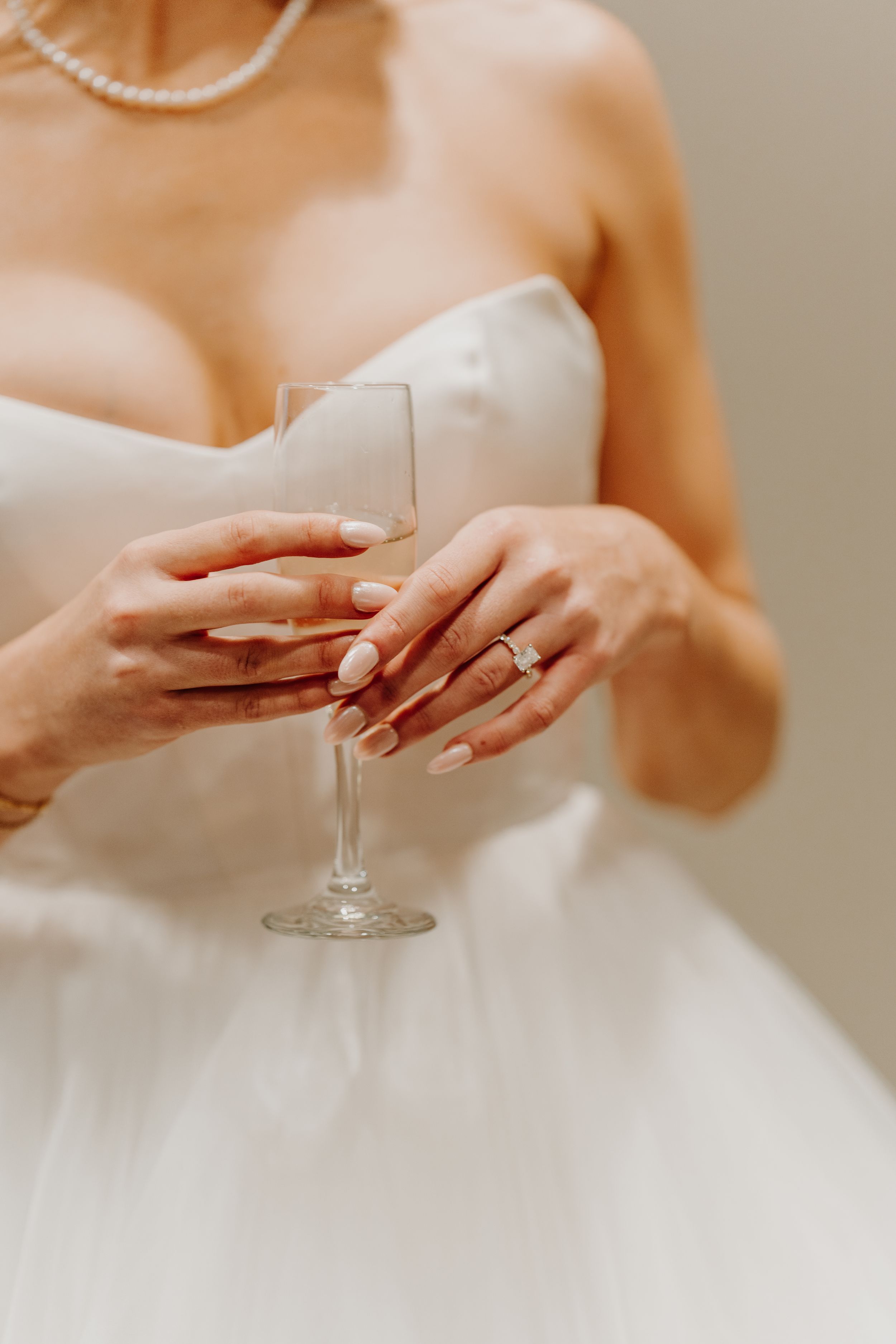 Close-up of bride's hands holding champagne glass, showing engagement ring and white dress details.