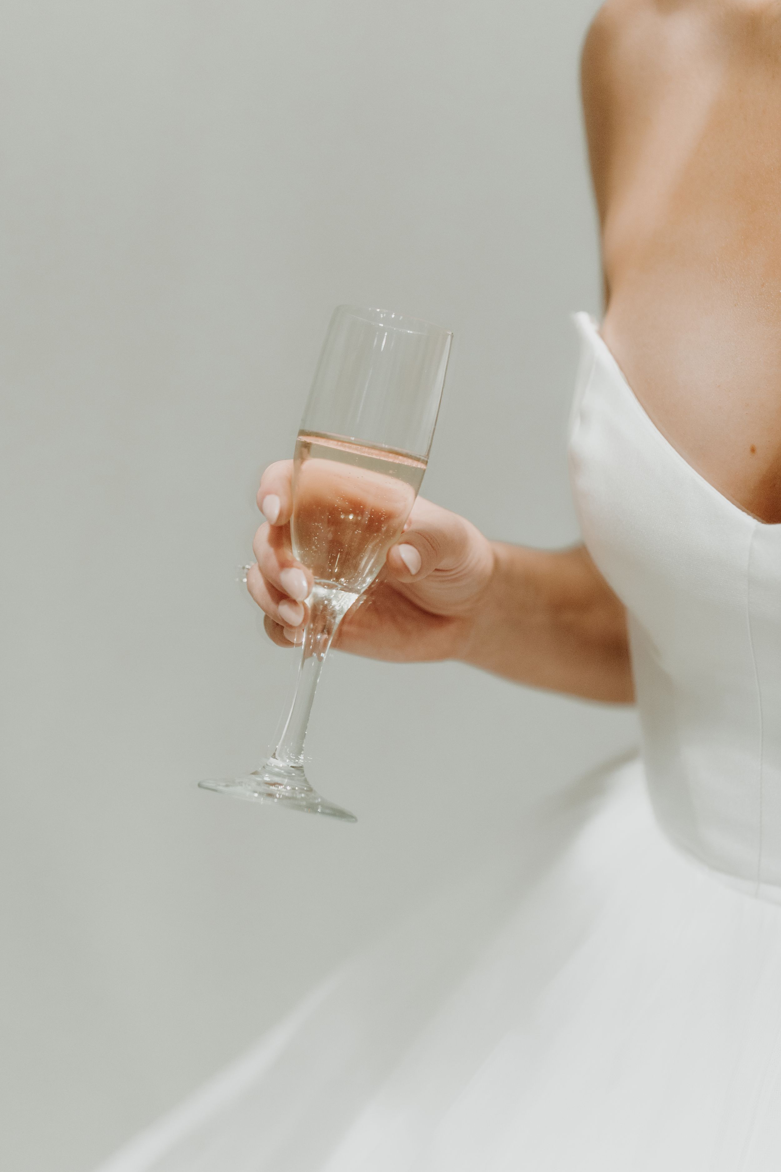 Bride in white dress holding champagne glass against light background.