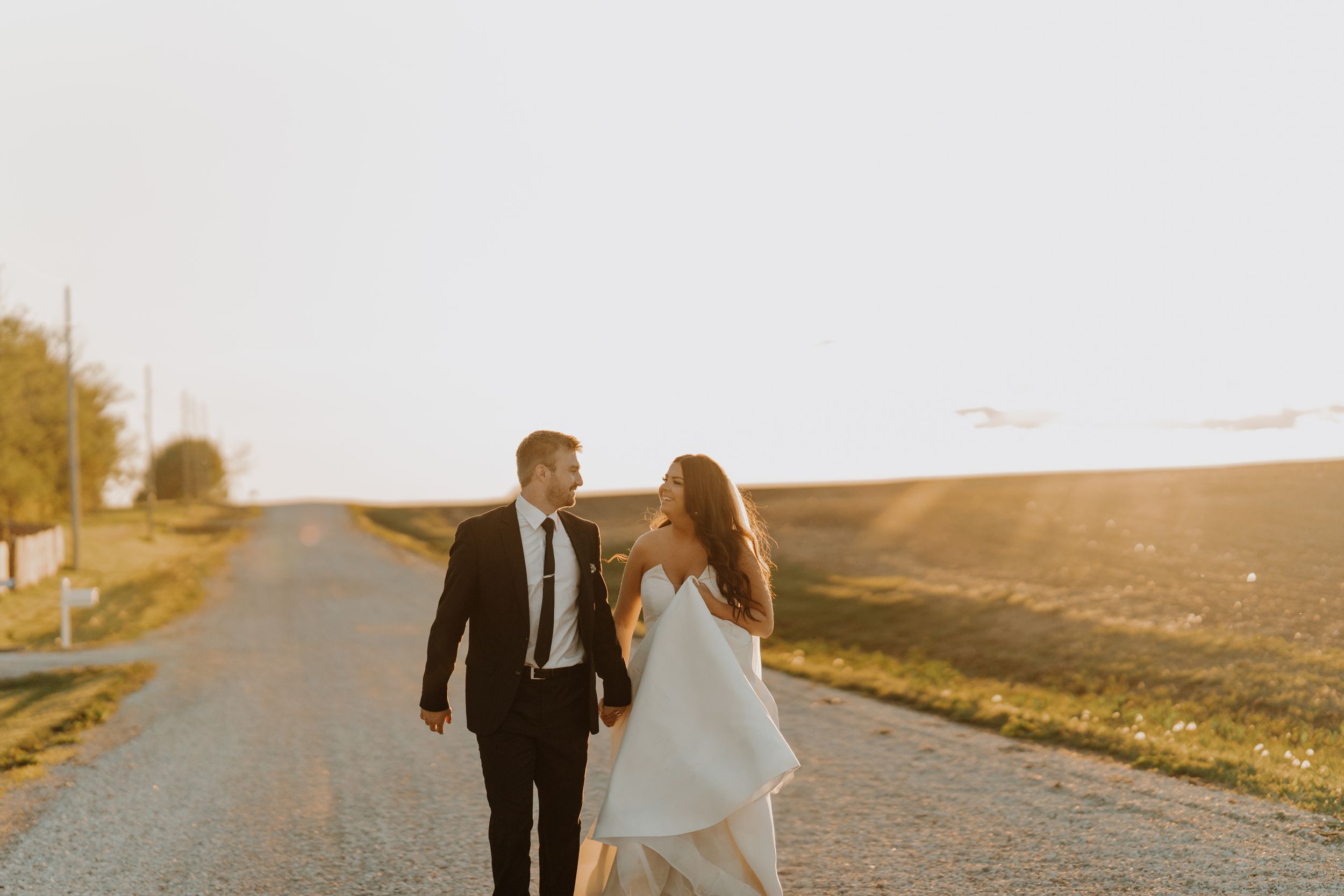 Bride and groom walking together on country road at sunset in wedding attire.