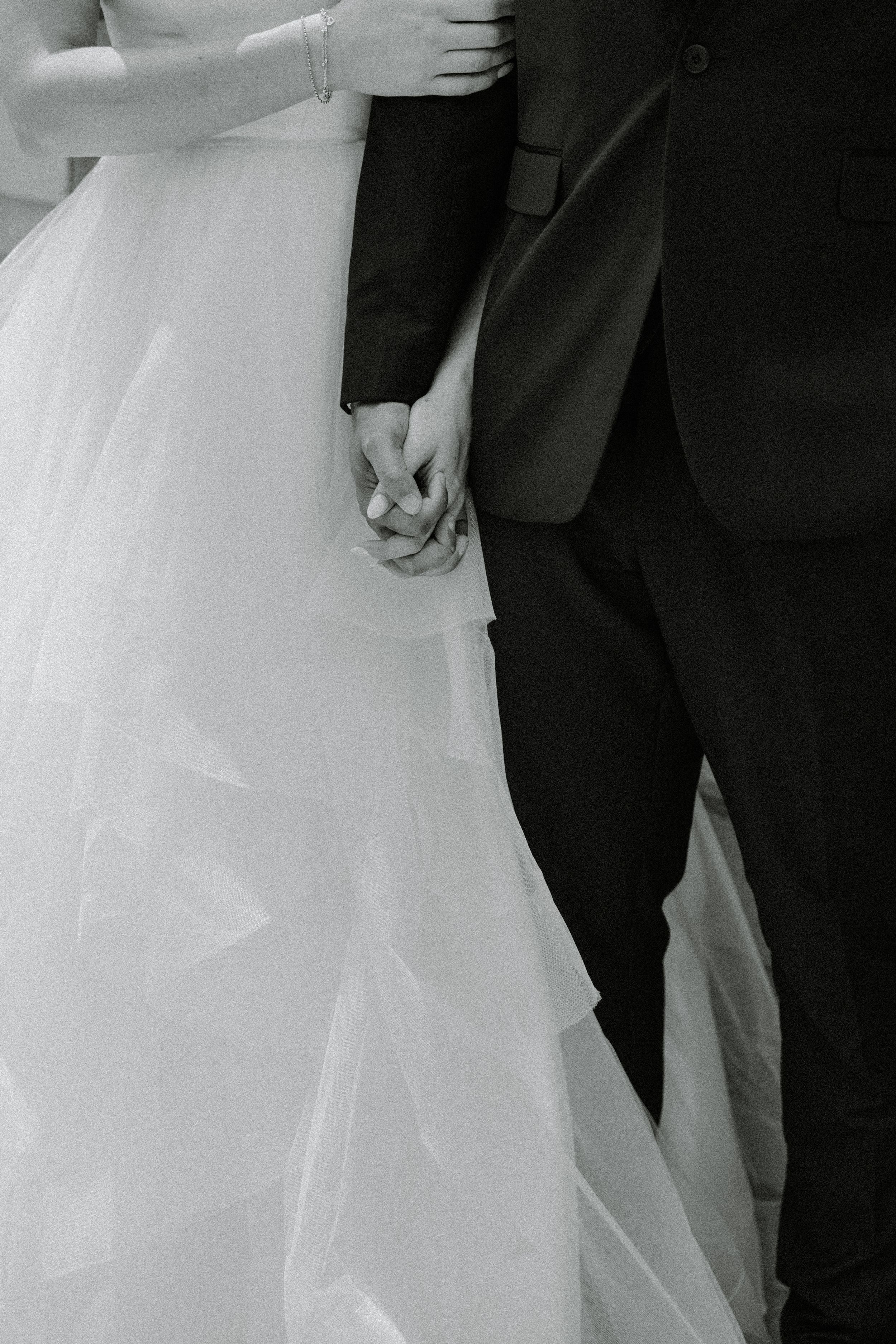 Black and white photo of bride and groom holding hands, showing dress and suit details.