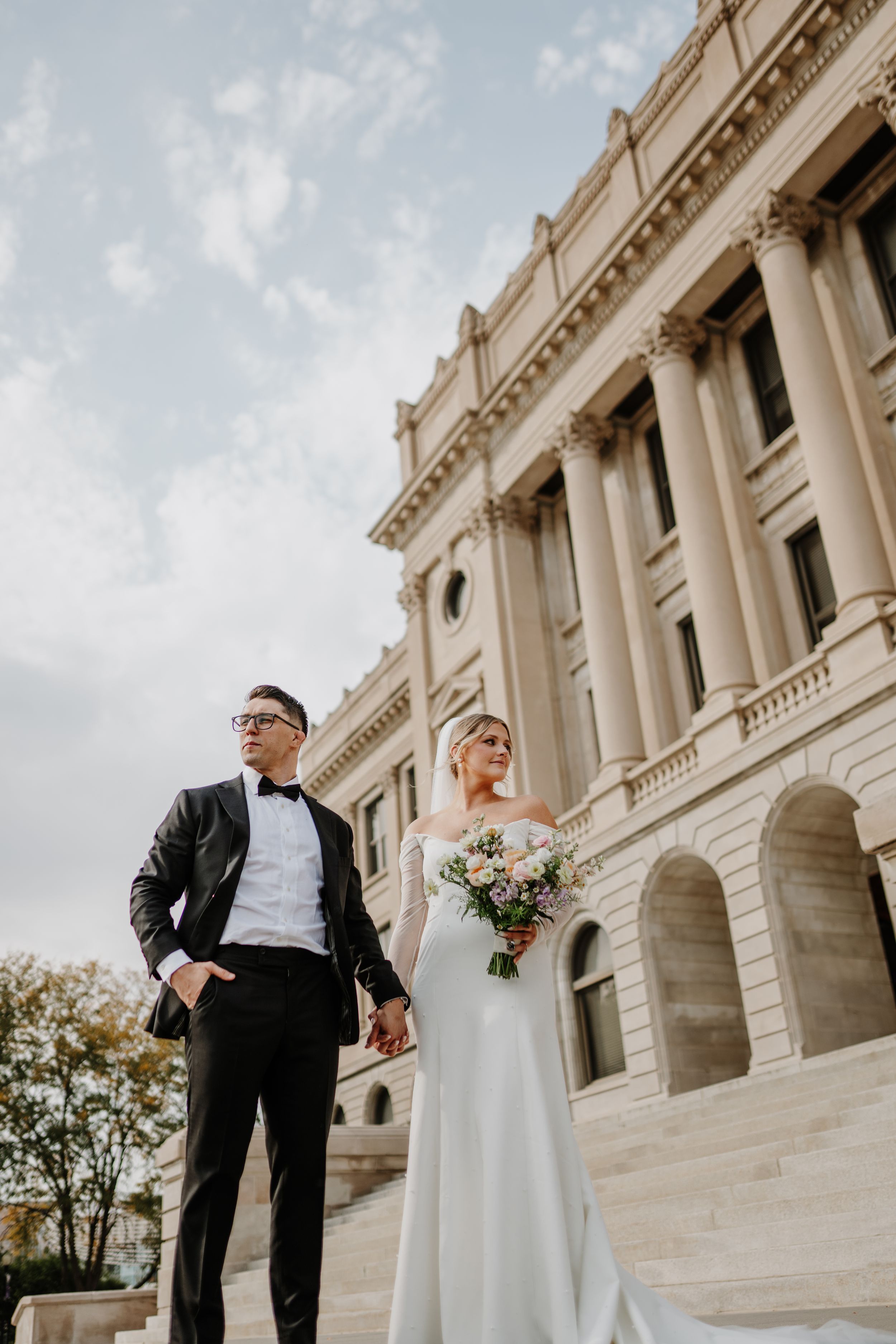 A wedding couple poses elegantly on grand stone steps of a neoclassical building with tall columns.