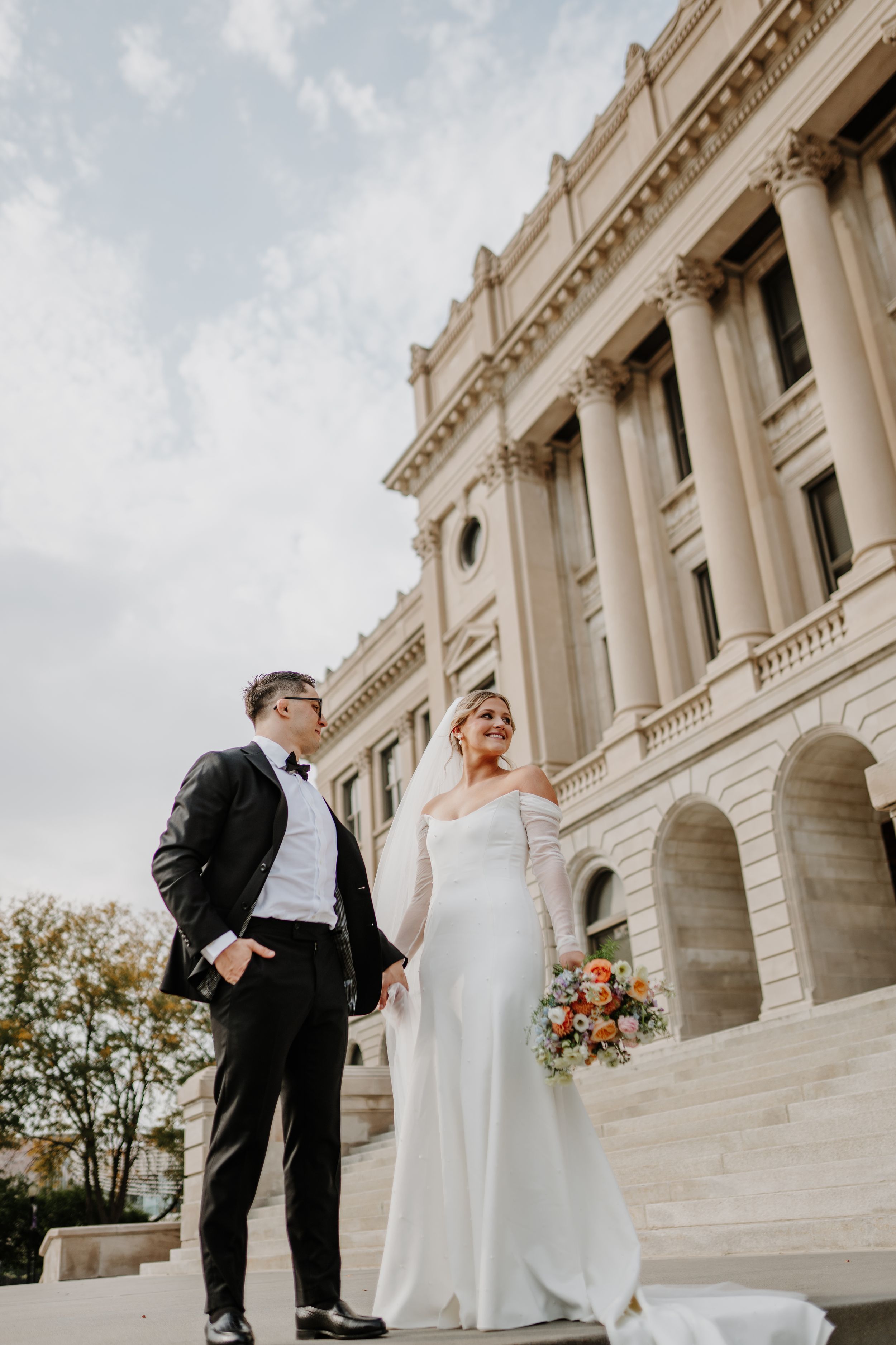 Newlyweds stand together on courthouse steps with bride holding colorful bouquet against classic architecture.