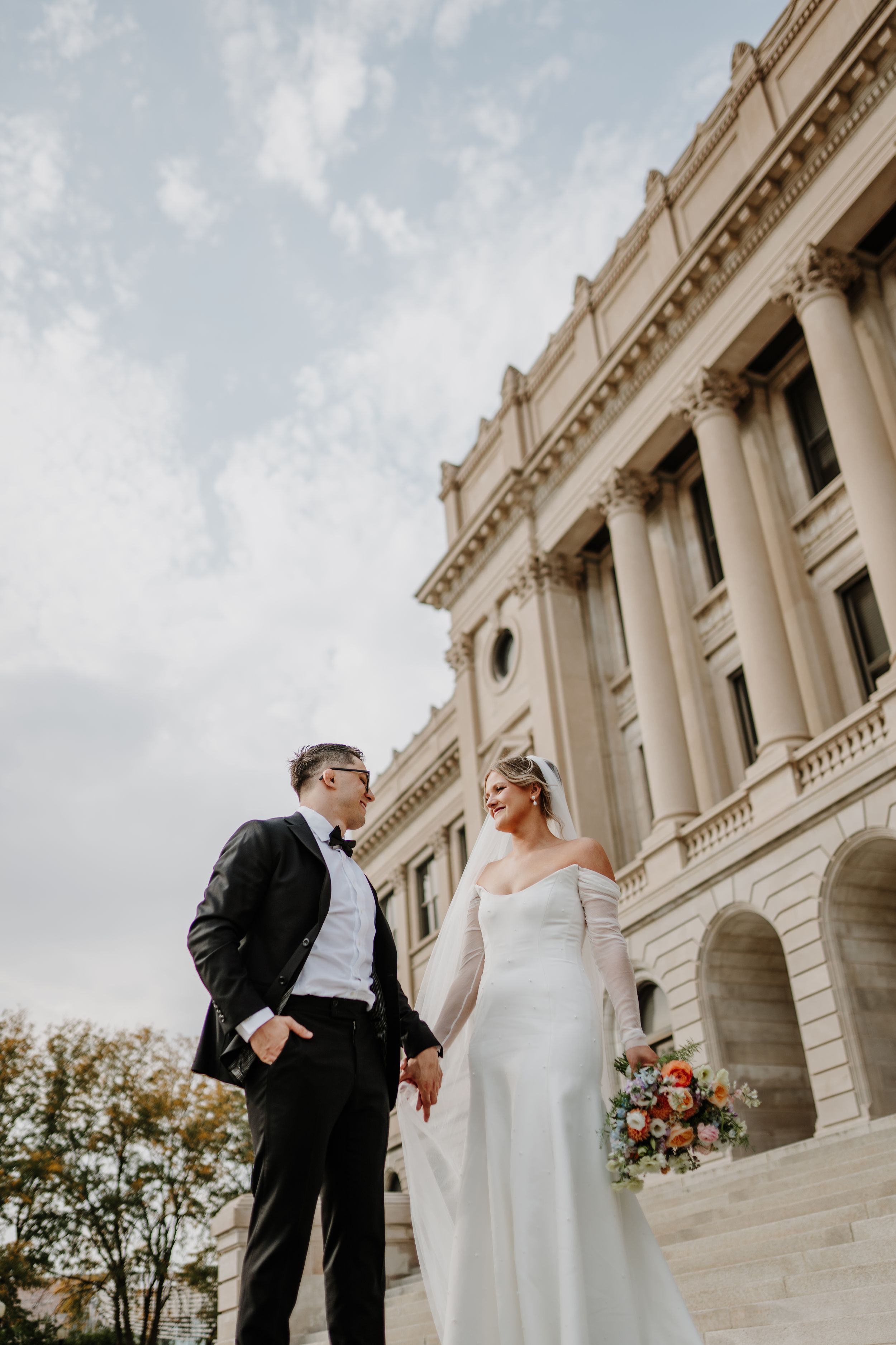 A romantic wedding portrait featuring couple in formal attire against ornate government building backdrop.