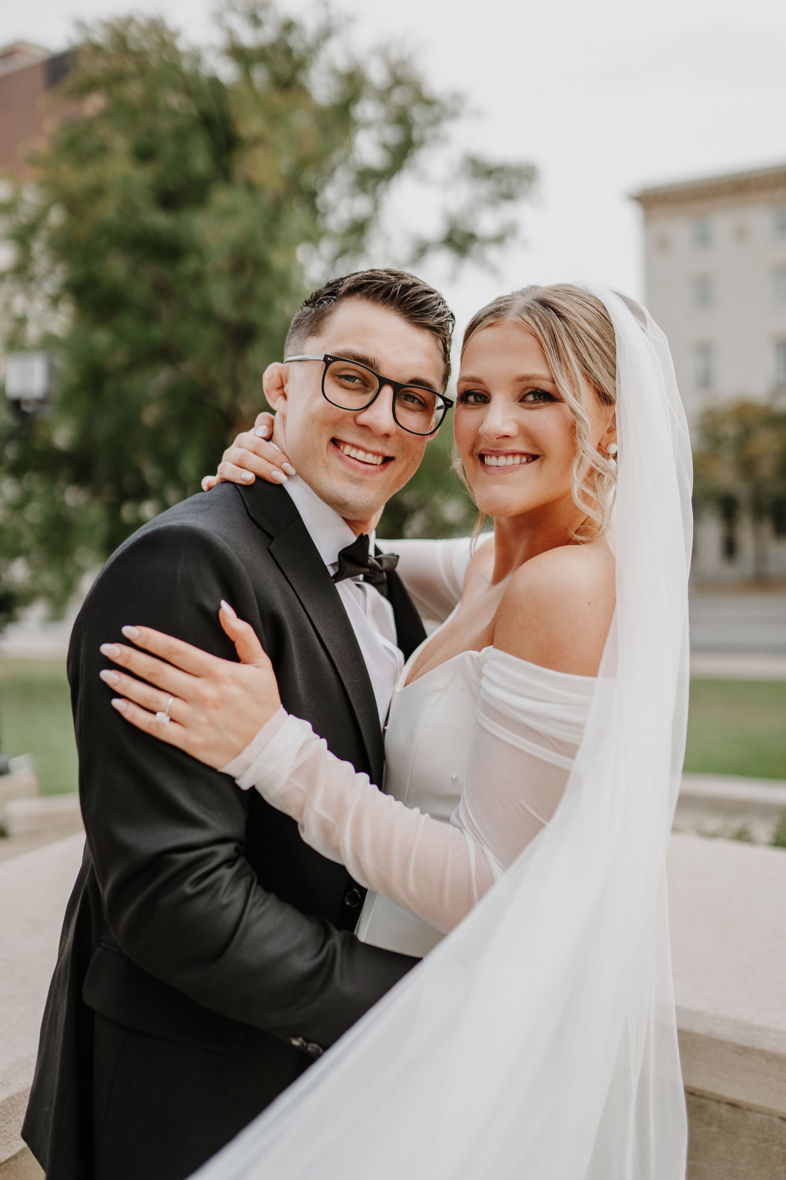 Joyful bride and groom embrace while showing off wedding attire in outdoor portrait.