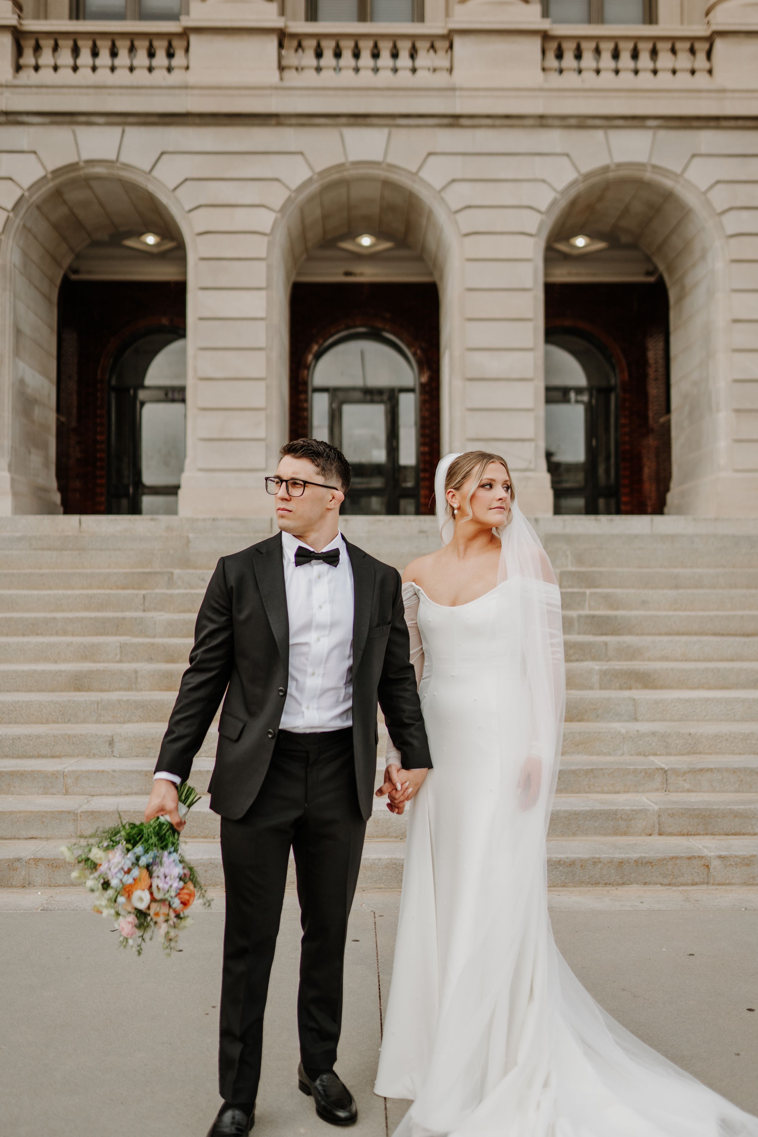 Stylish bride and groom descend courthouse steps while holding vibrant floral bouquet.