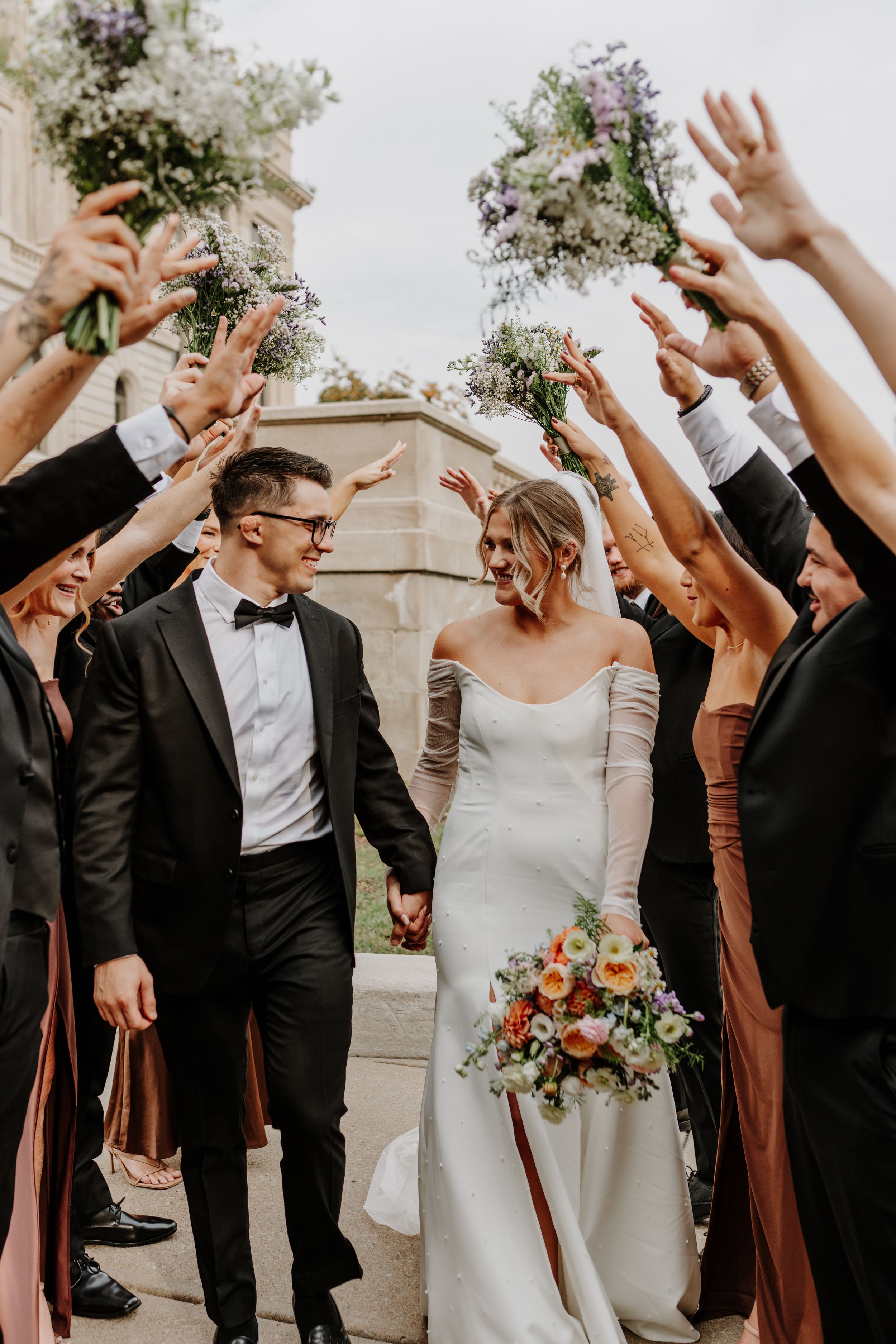 Wedding party in brown dresses and black tuxedos celebrating with raised arms and joyful expressions.