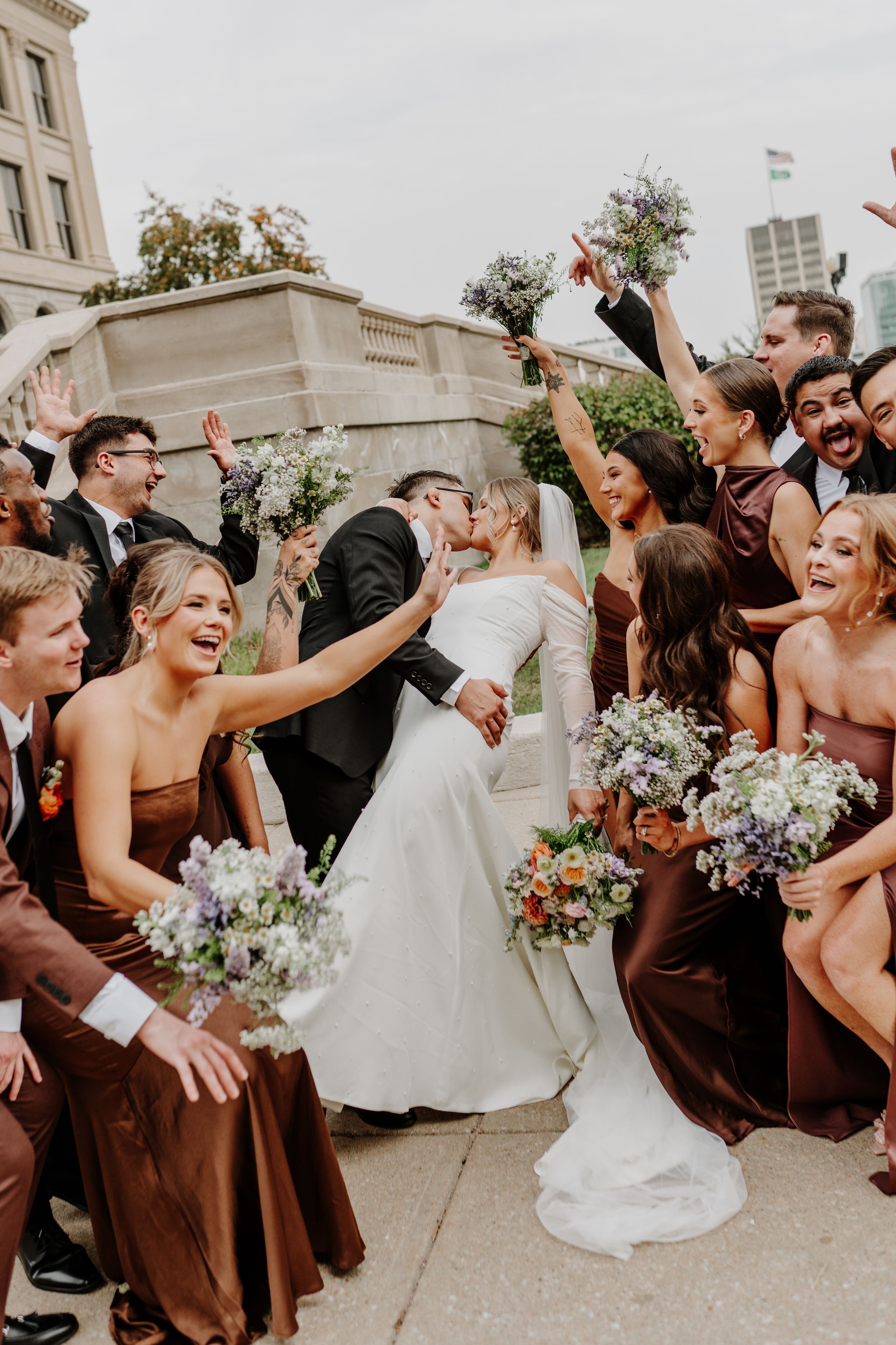 Energetic wedding party celebrates with raised arms and excited expressions outside a light-colored building.