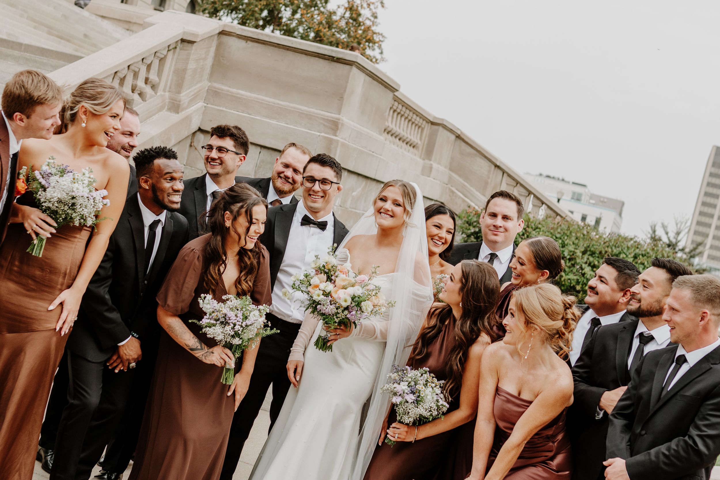 Wedding party in brown dresses and formal attire poses against a modern building facade.