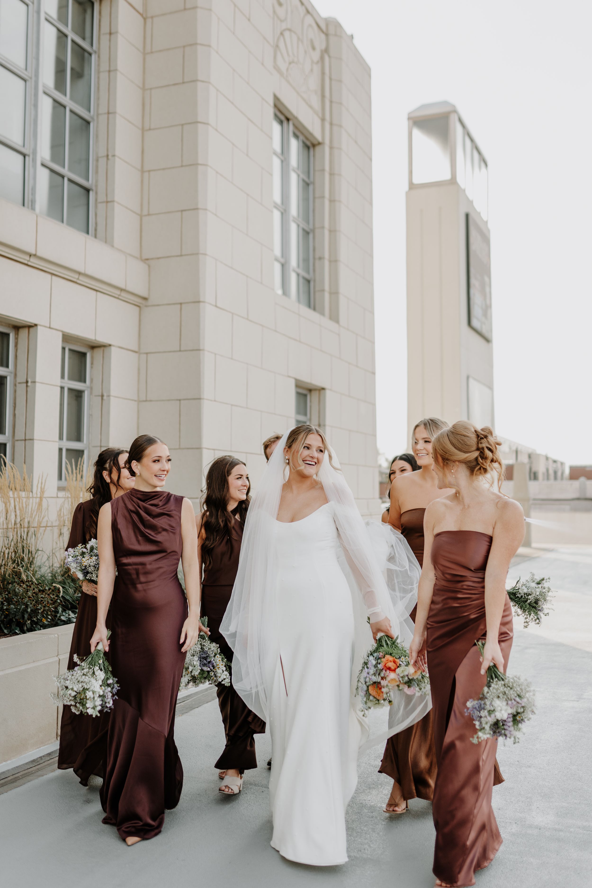 Wedding party in brown bridesmaid dresses and white gown walking together against light stone building.