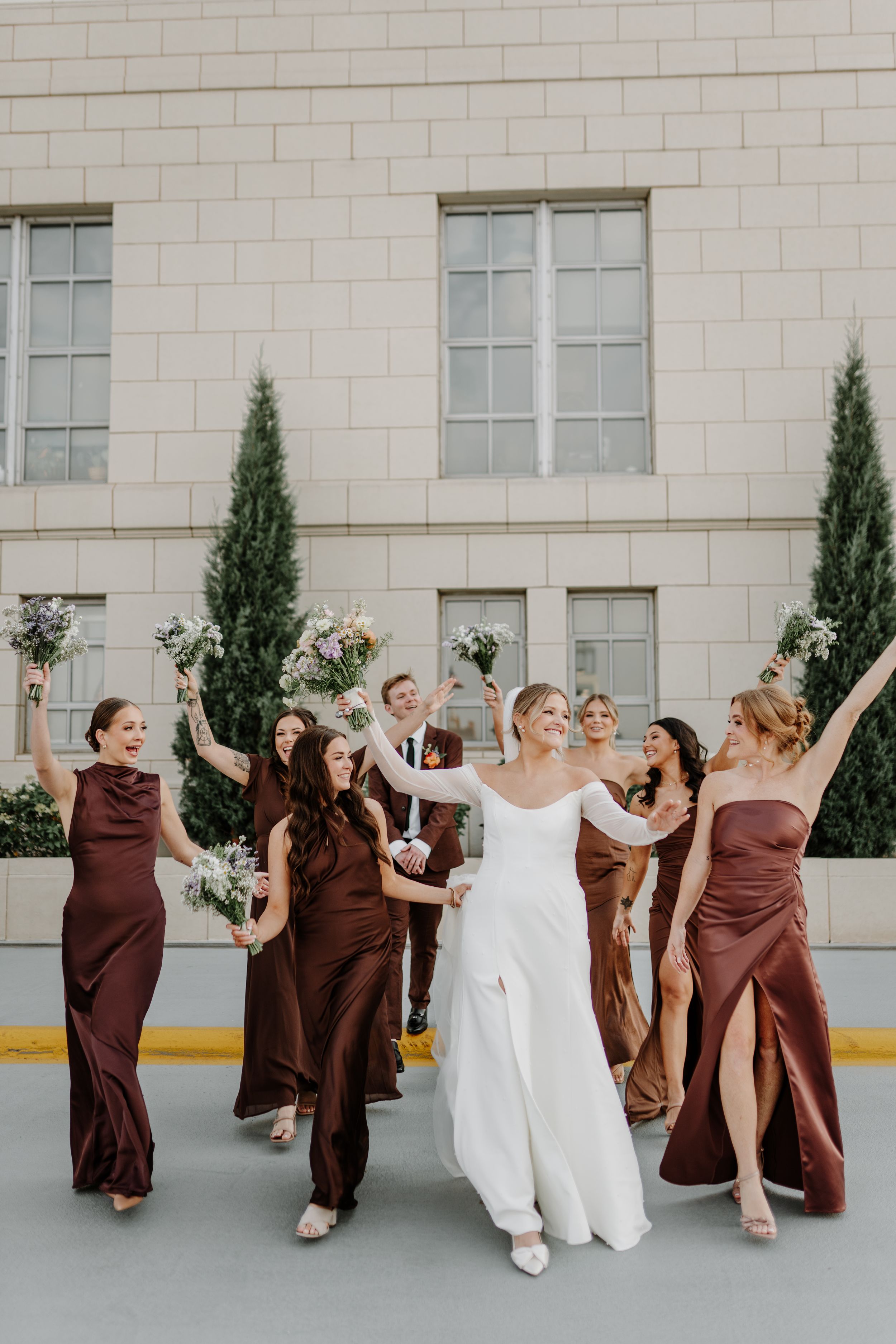Fun wedding party photo with bridesmaids in brown dresses posing playfully against white building.
