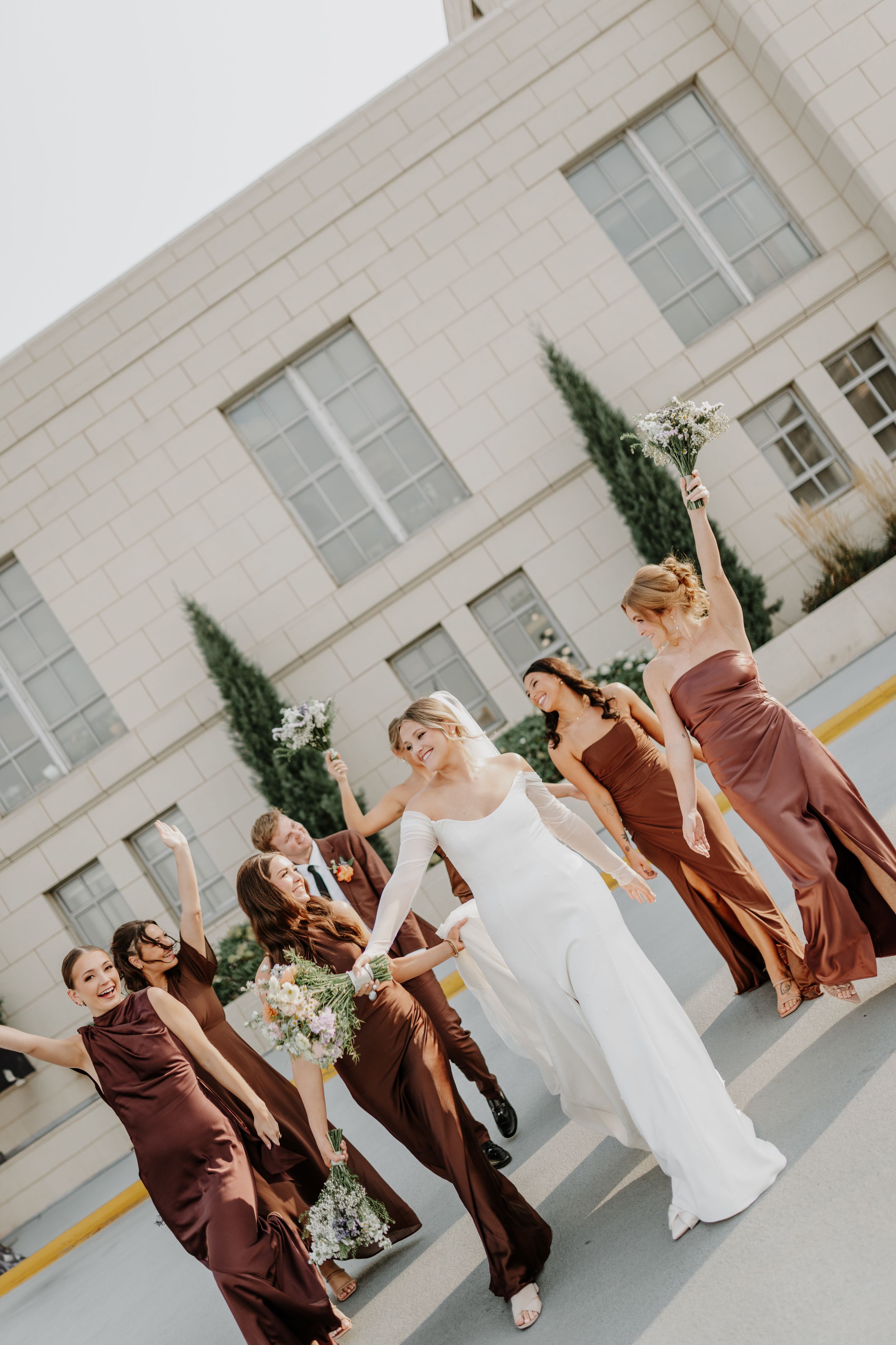 Candid shot of bridesmaids in brown dresses celebrating with the bride against a modern building.