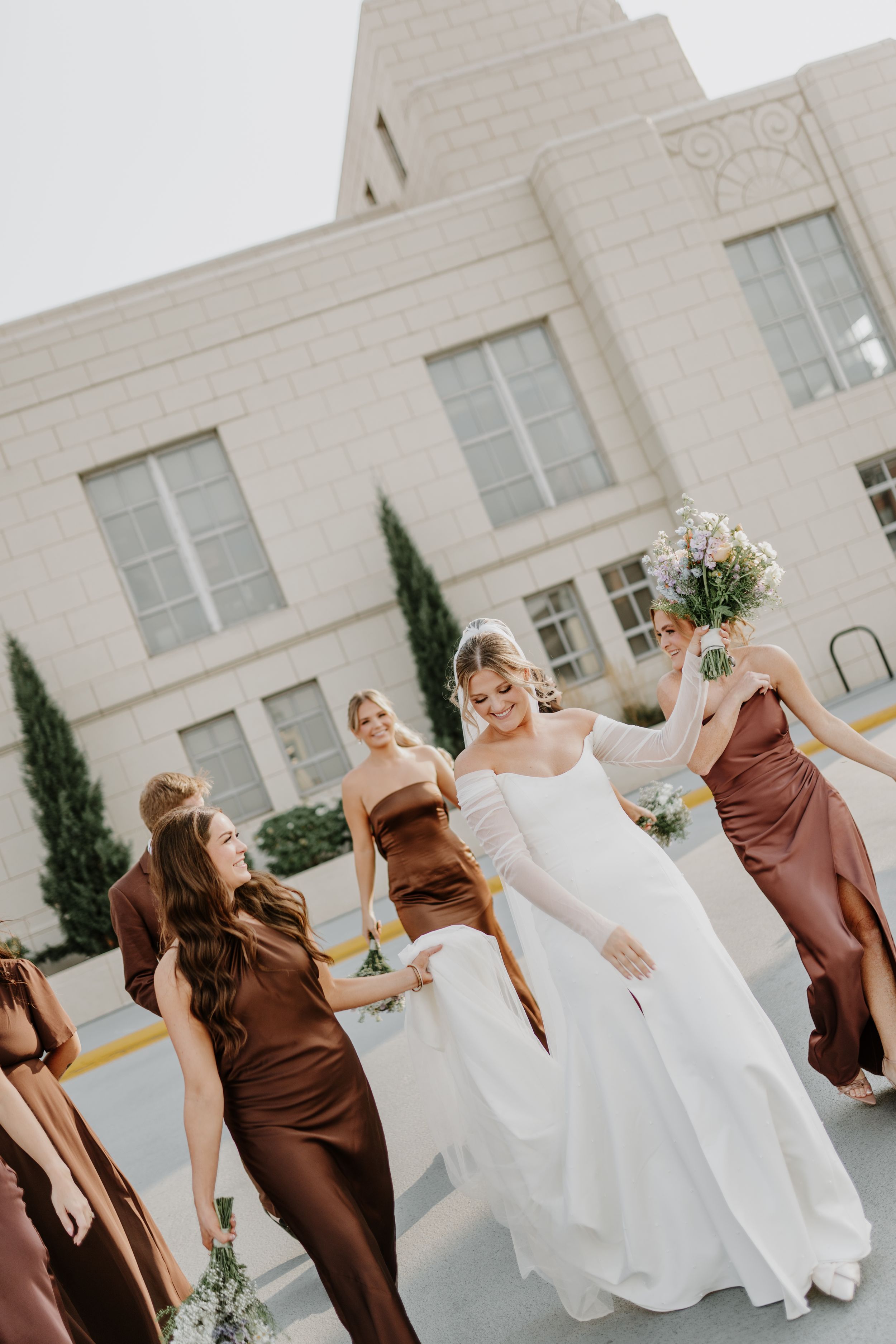 Wedding party dancing and celebrating outdoors against a light stone building backdrop.