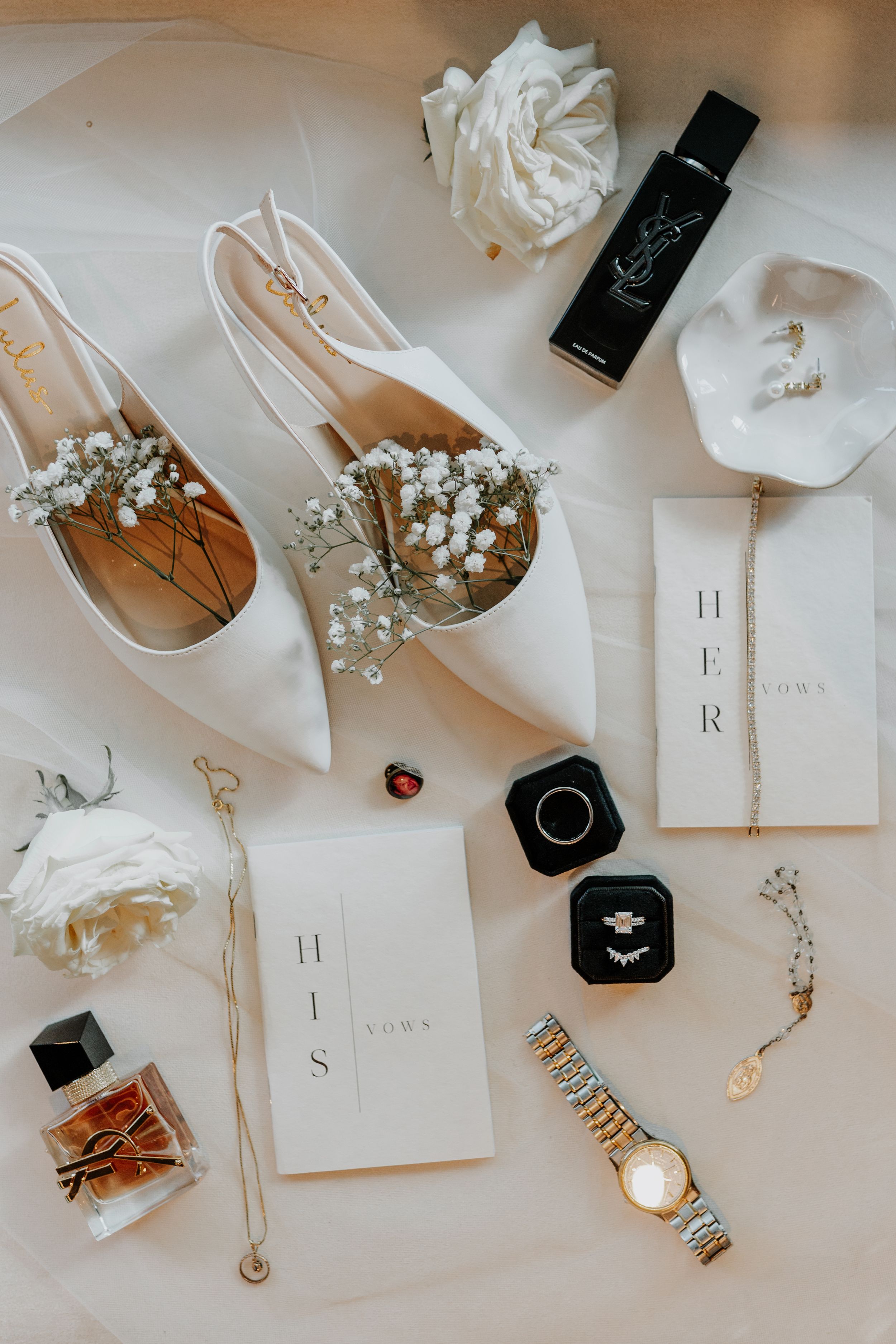 Wedding accessories including shoes, rings, and invitations arranged with white roses.