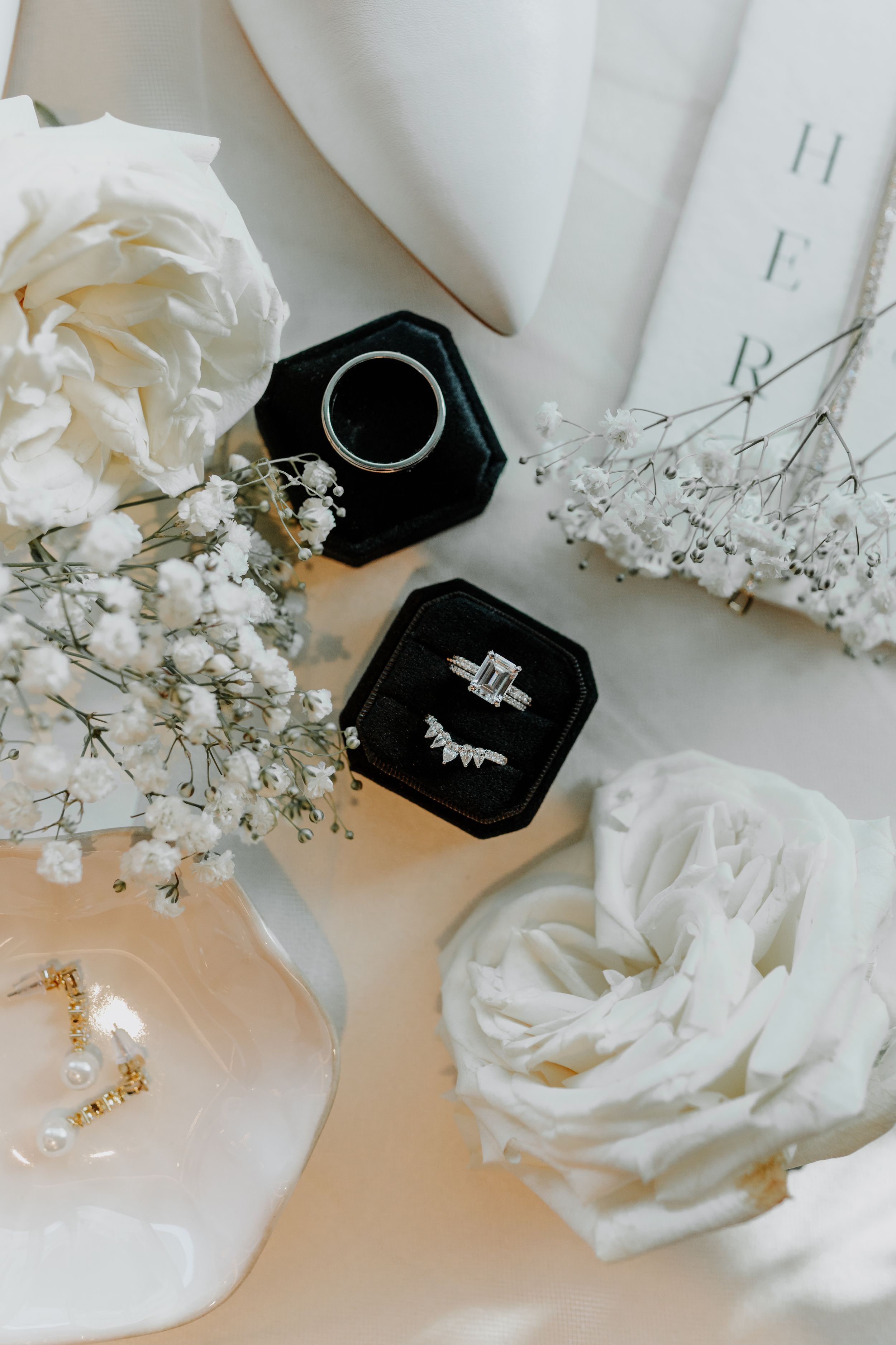 Wedding details flatlay with white roses, jewelry, and black ring boxes.