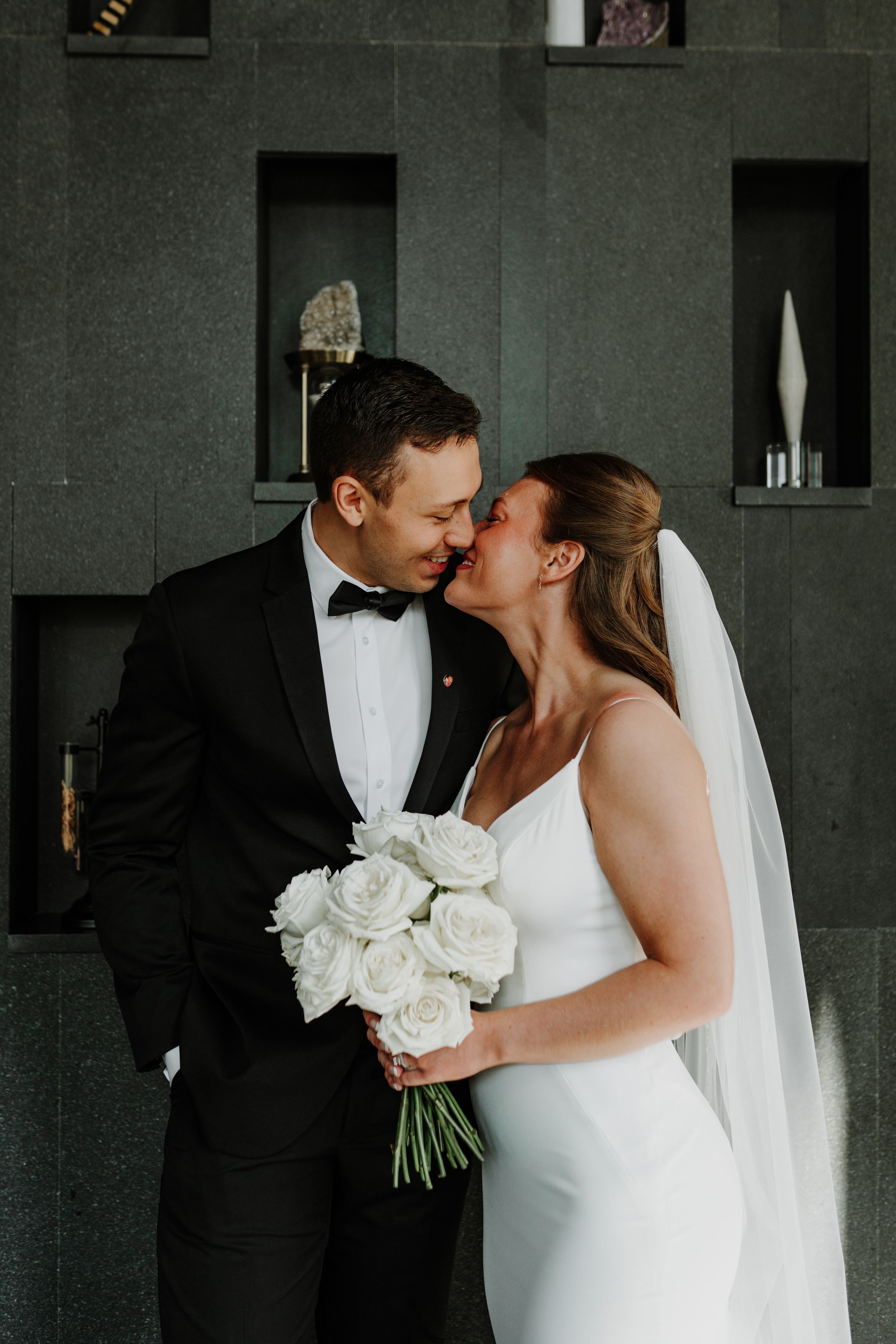 Bride holding white roses embraces groom in front of modern grey wall with display niches.