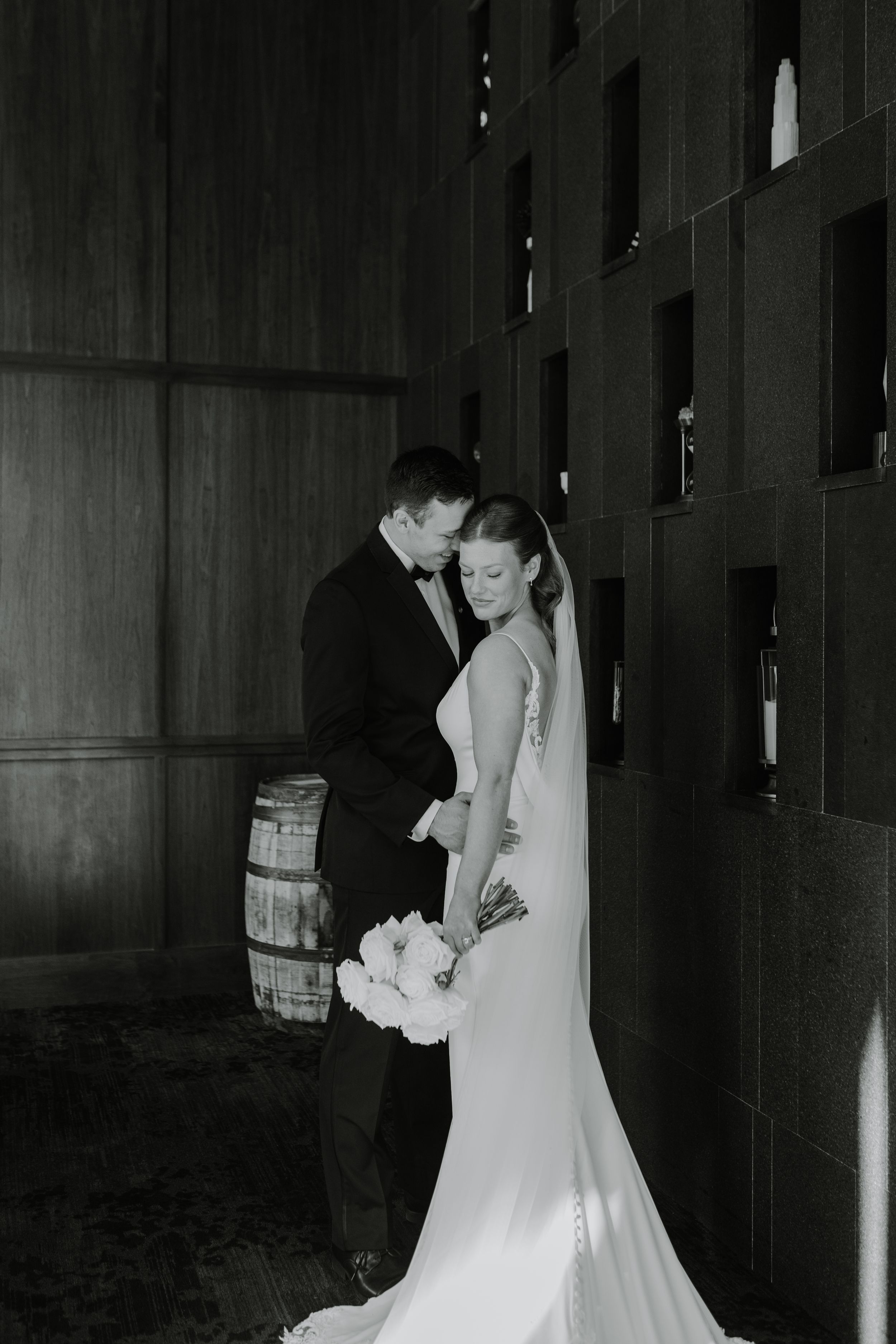 Black and white wedding portrait shows couple in tender moment by rustic wooden barrel.